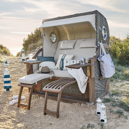Sillón de playa de mimbre con cojines beige y reposapiés de madera en la arena, ideal para relajarse