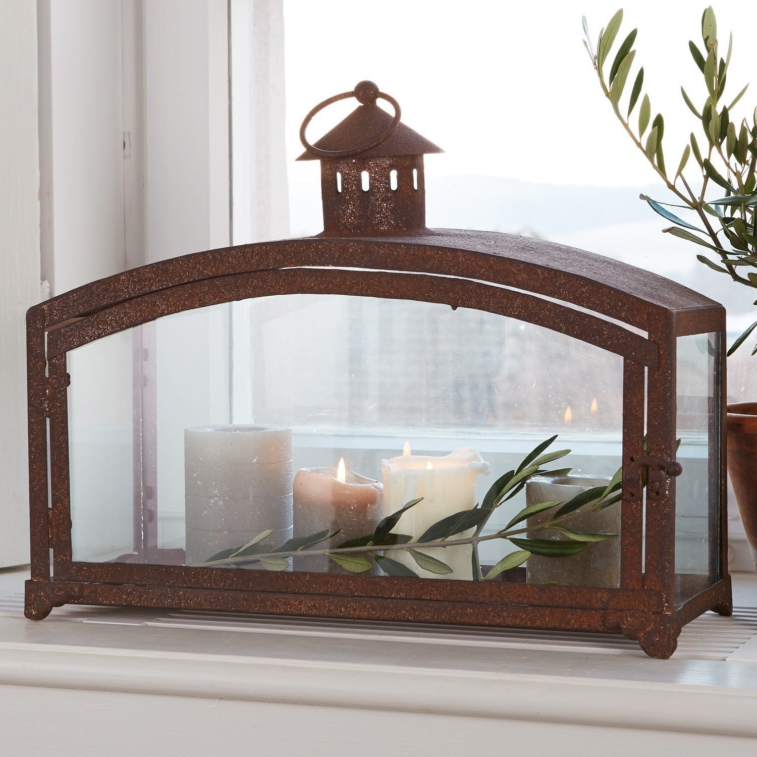 Portavelas Miora Linterna de metal oxidado con paneles de vidrio, velas encendidas y ramas verdes decorativas