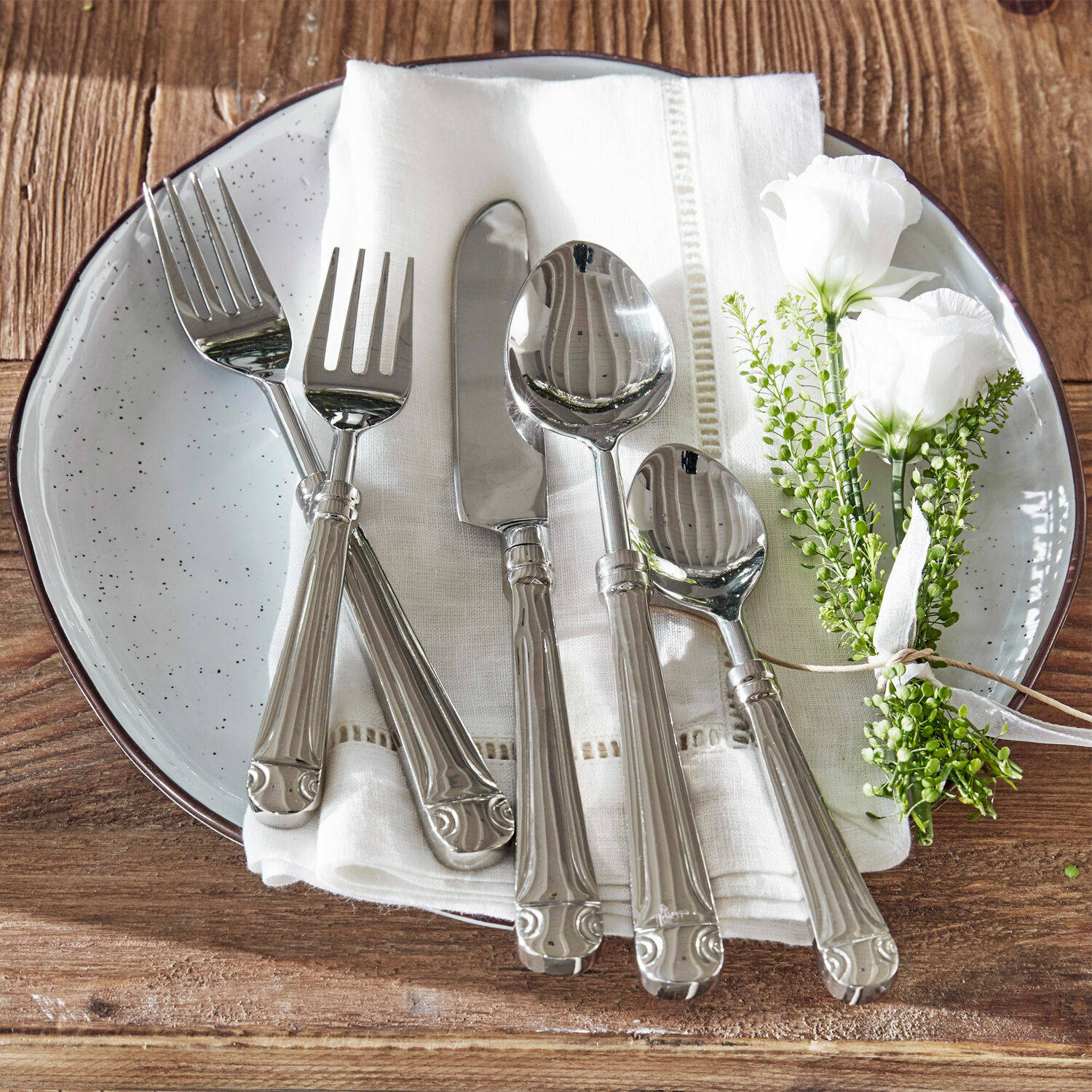 Cubiertos Eriat Juego de cubiertos plateados sobre servilleta blanca con decoración floral en mesa de madera