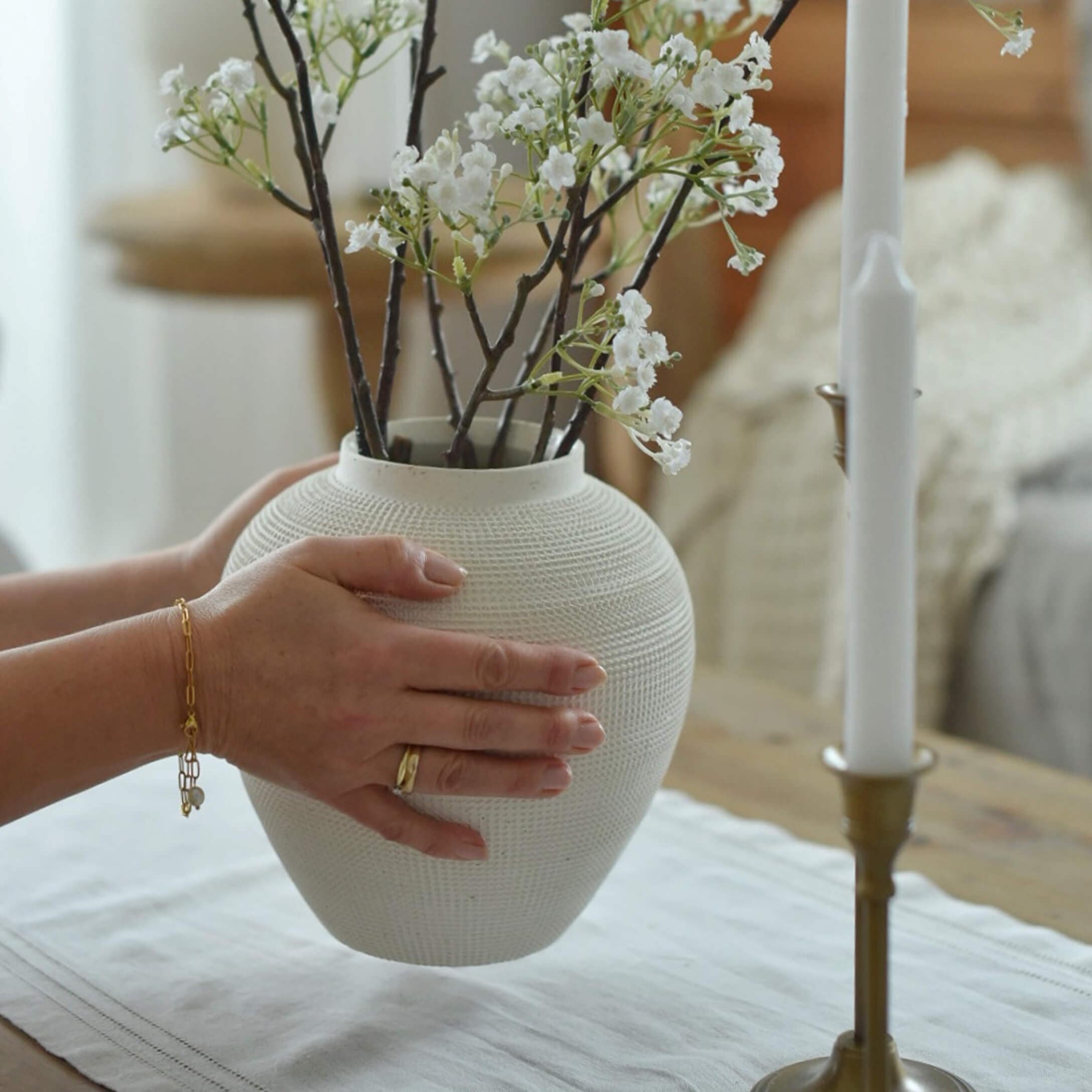 Jarrón de cerámica blanca con textura sostenido por manos con ramas de flores blancas sobre mesa
