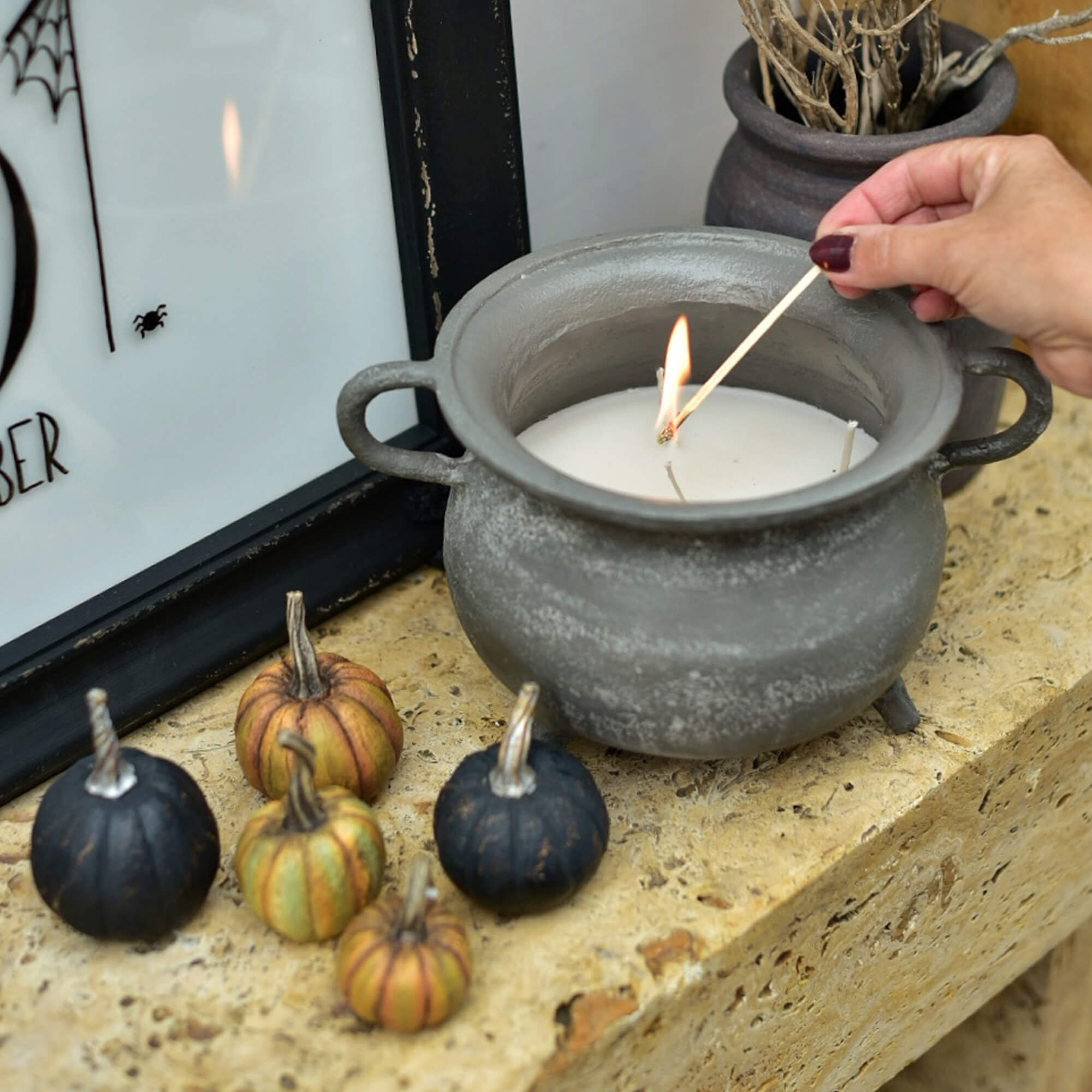 Vela de cerámica gris en forma de caldero con cera blanca y mini calabazas decorativas sobre repisa de piedra
