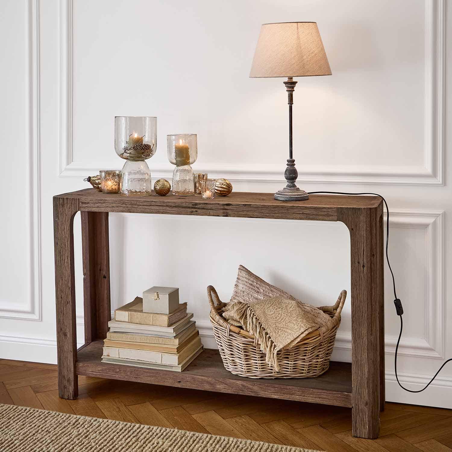 Consola de madera con lámpara de mesa, velas en portavelas de cristal y cesta de mimbre con textiles