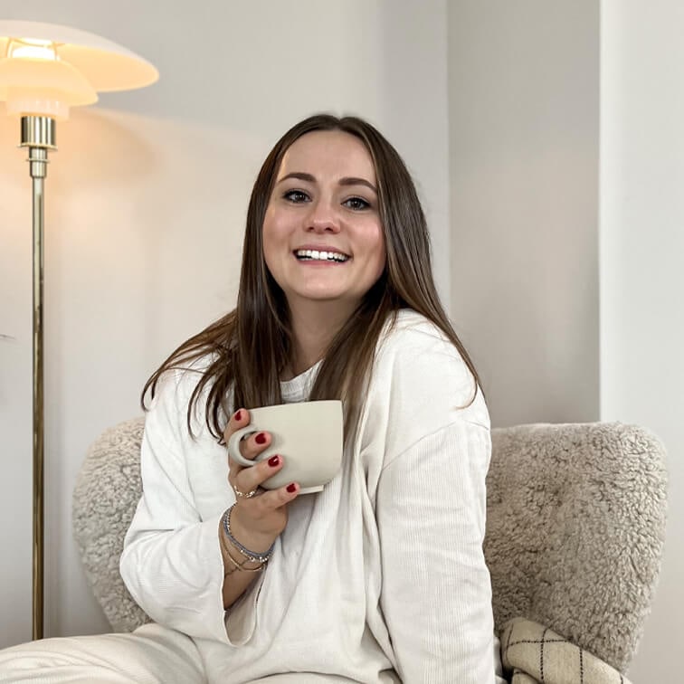 Mujer sonriente con suéter blanco sosteniendo una taza beige sentada en un sillón cómodo en un interior acogedor