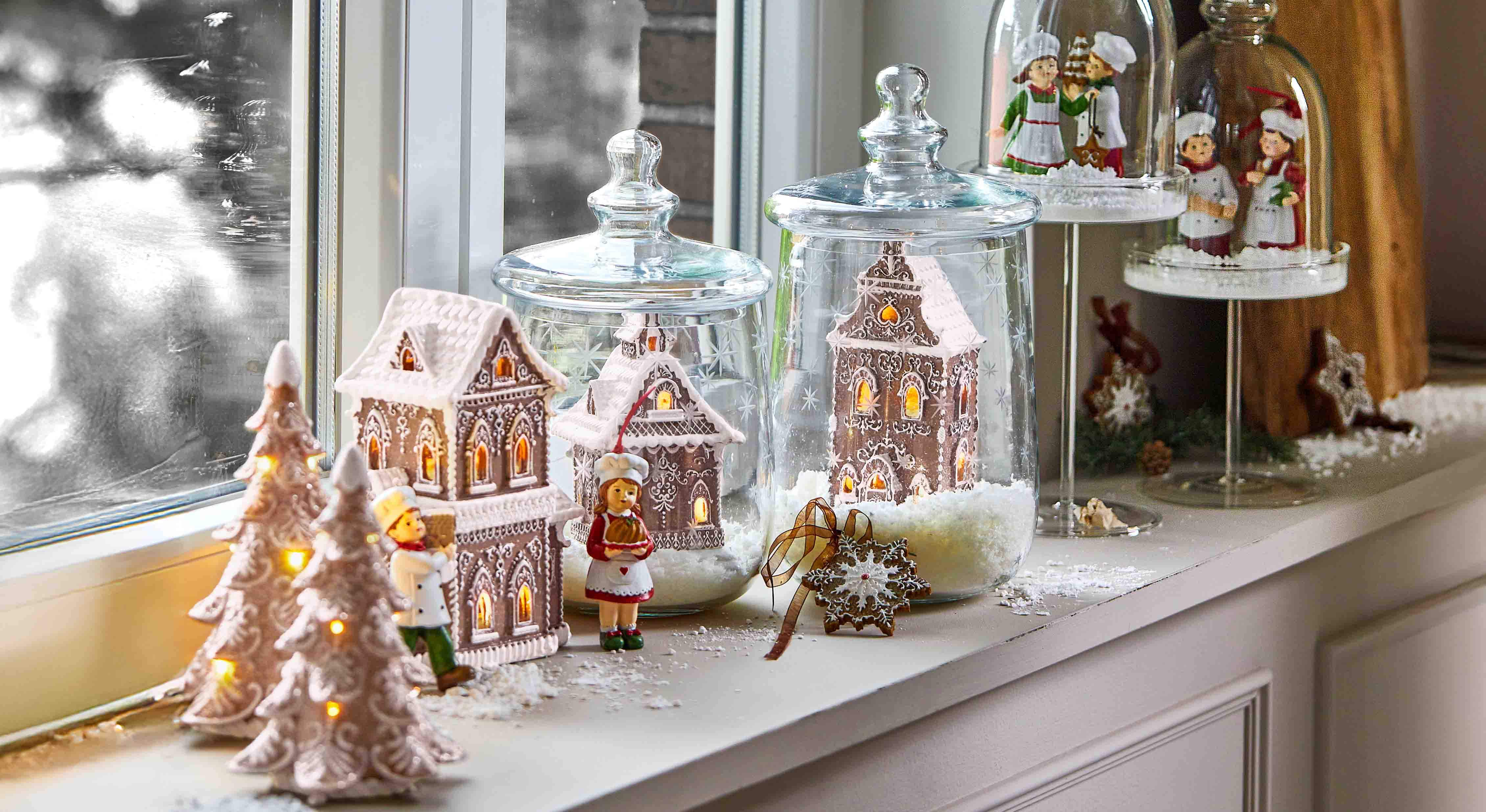 Casitas de jengibre decorativas y figuras de cocineros bajo campanas de cristal en el alféizar con luces invernales