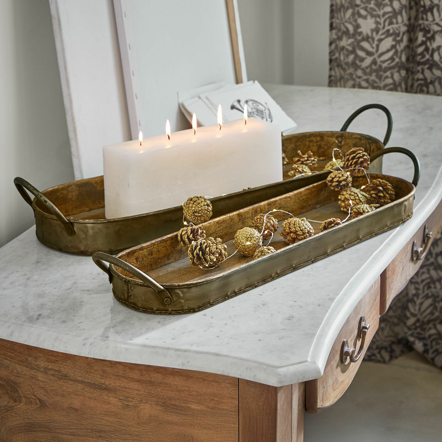 Set de 2 bandejas Célianne Bandeja decorativa metálica dorada sobre encimera de mármol con vela blanca y adornos de piñas