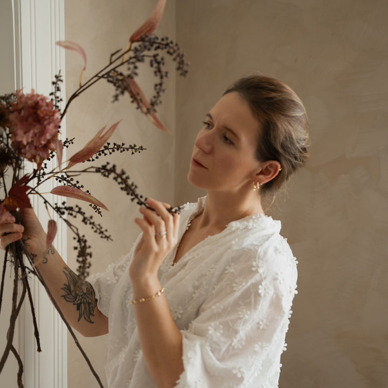 Mujer con blusa blanca de encaje arreglando decoración floral seca en interior luminoso