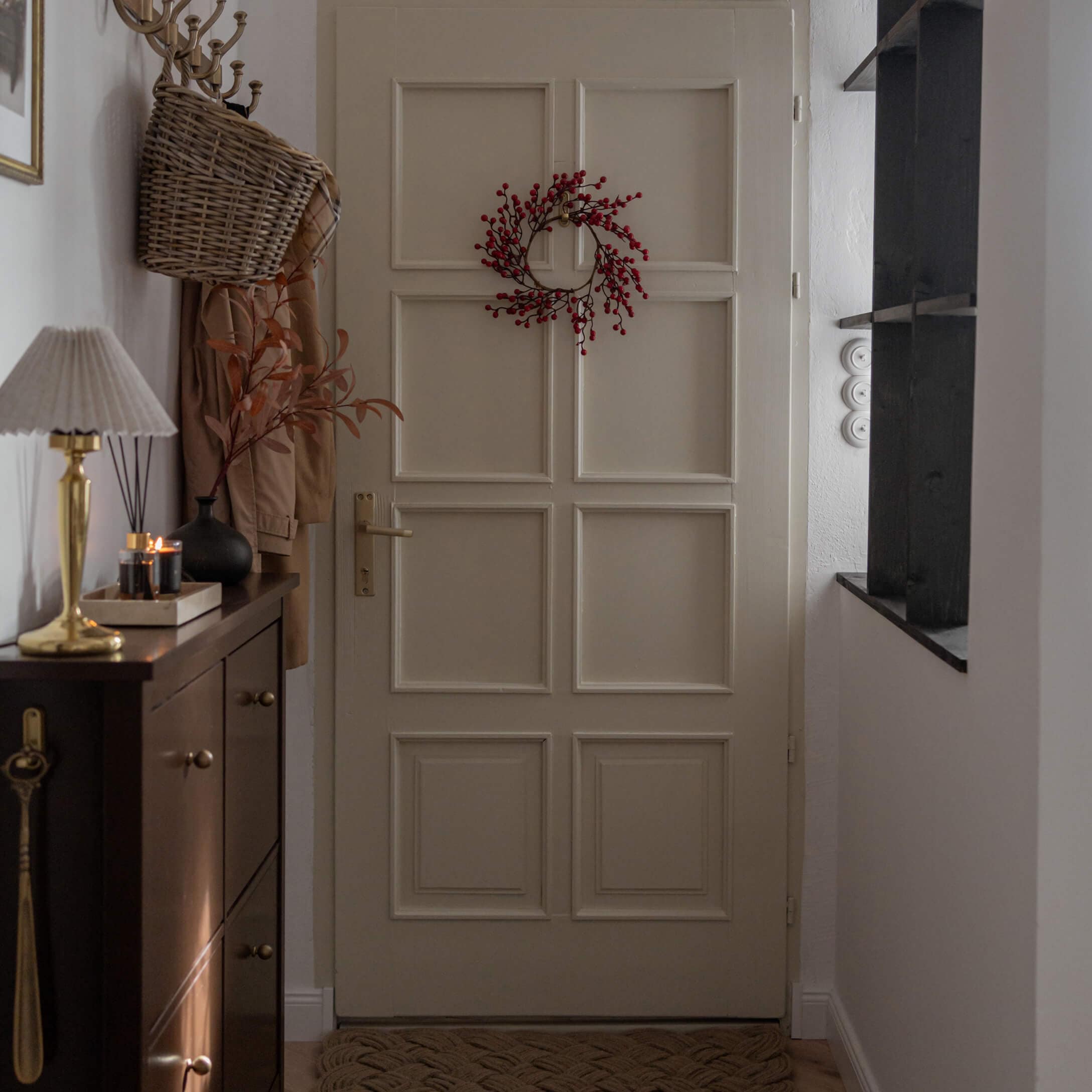 Recibidor con puerta beige decorada con corona roja y mueble oscuro con lámpara y velas encendidas