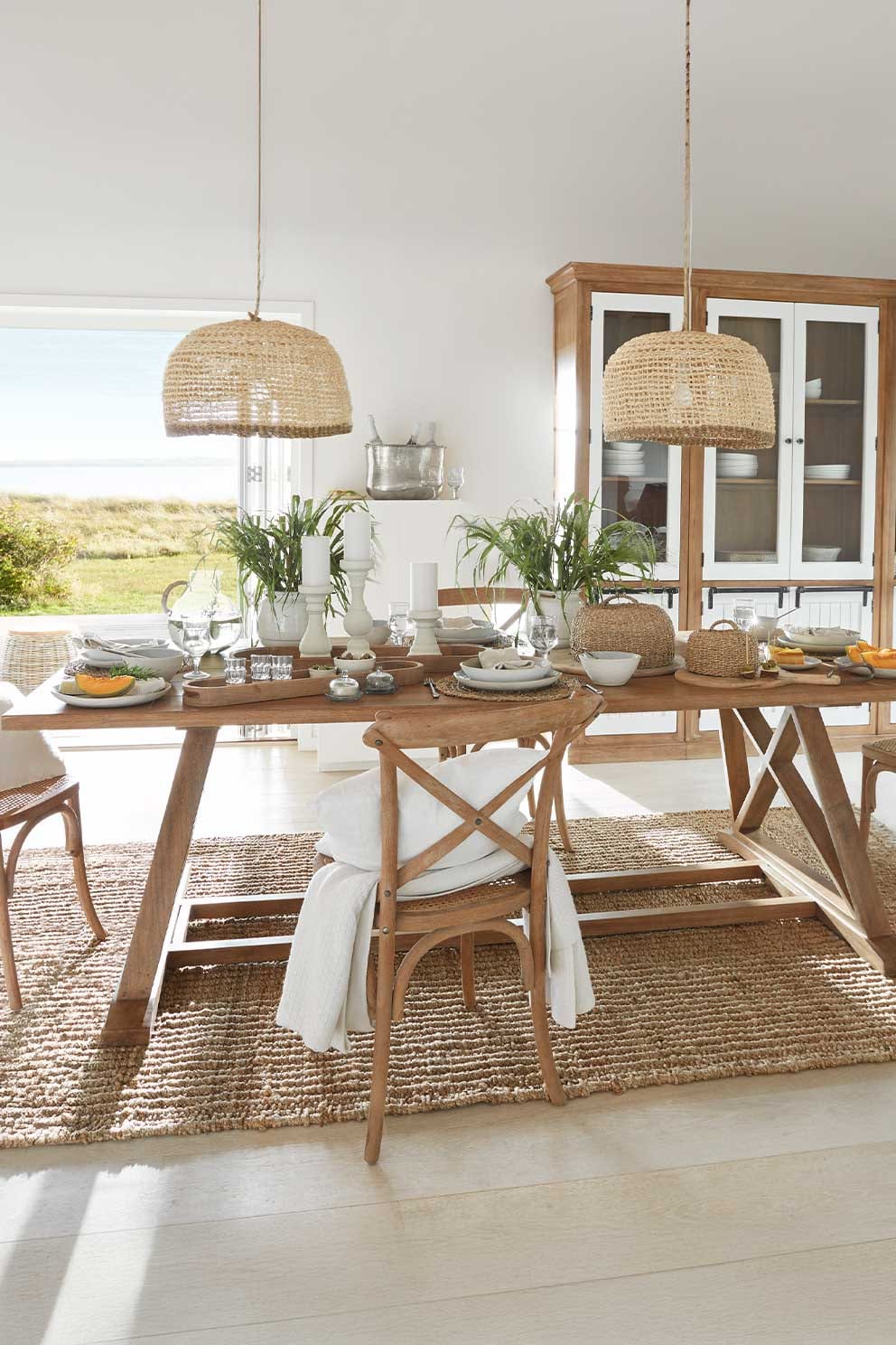 Comedor con mesa y sillas de madera, lámparas colgantes de mimbre natural, plantas decorativas y vajilla blanca