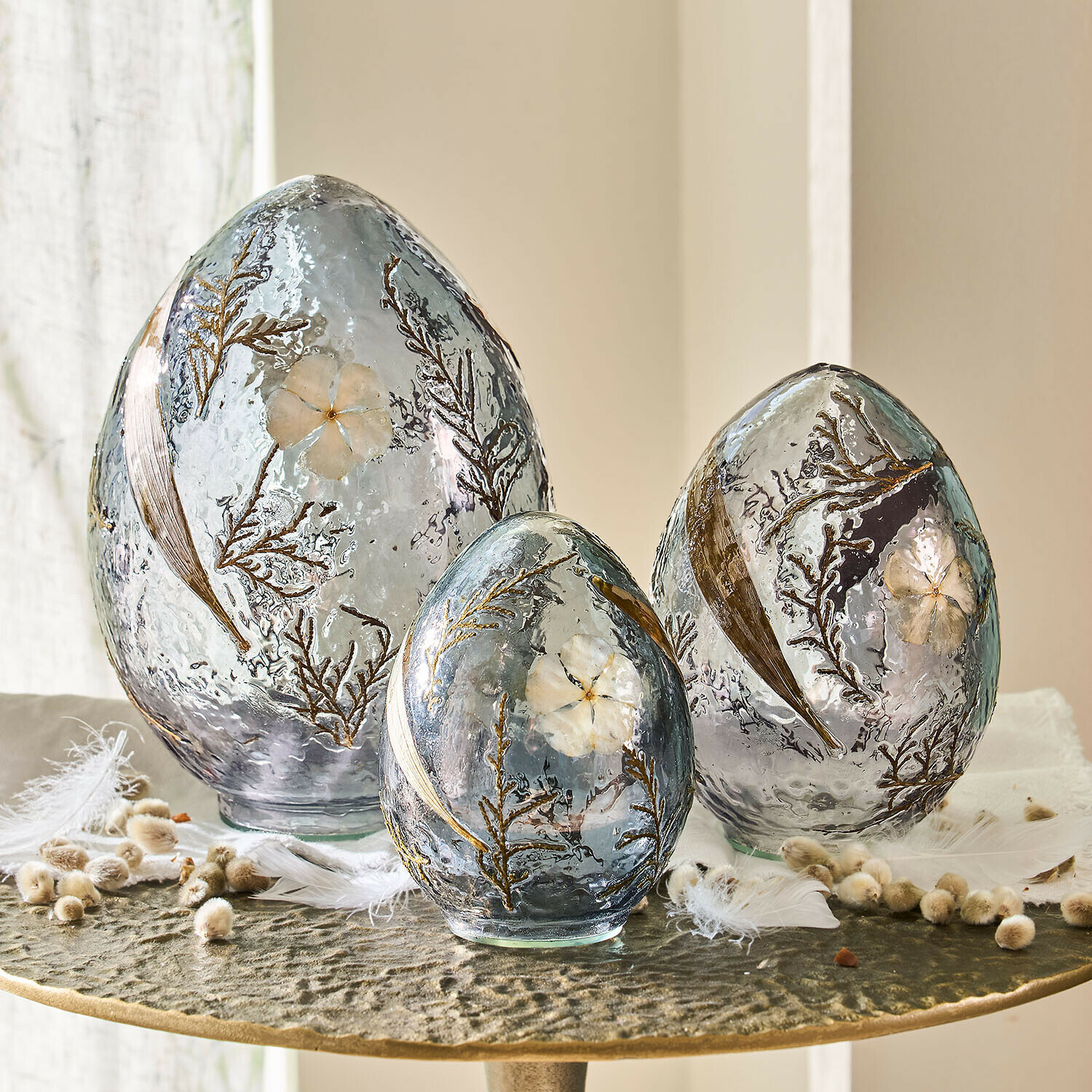 Set de 3 huevos decorativos Lunavelle Conjunto de tres huevos decorativos de vidrio gris con flores y hojas incrustadas sobre mesa