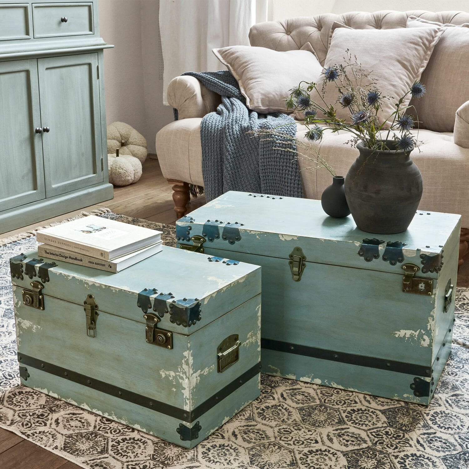 Set de 2 baúles Gorman Dos baúles de madera azul con herrajes metálicos sobre alfombra en sala con sofá beige