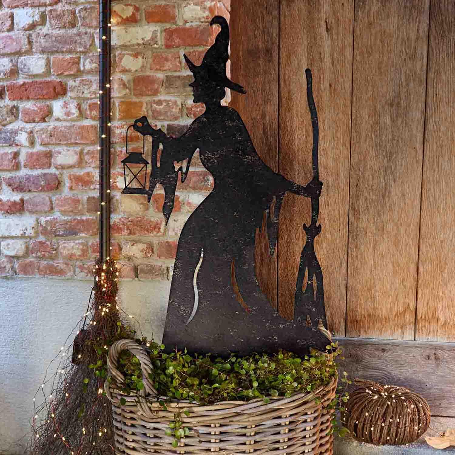 Decoración metálica de silueta de bruja con linterna y escoba en cesta de mimbre con plantas verdes