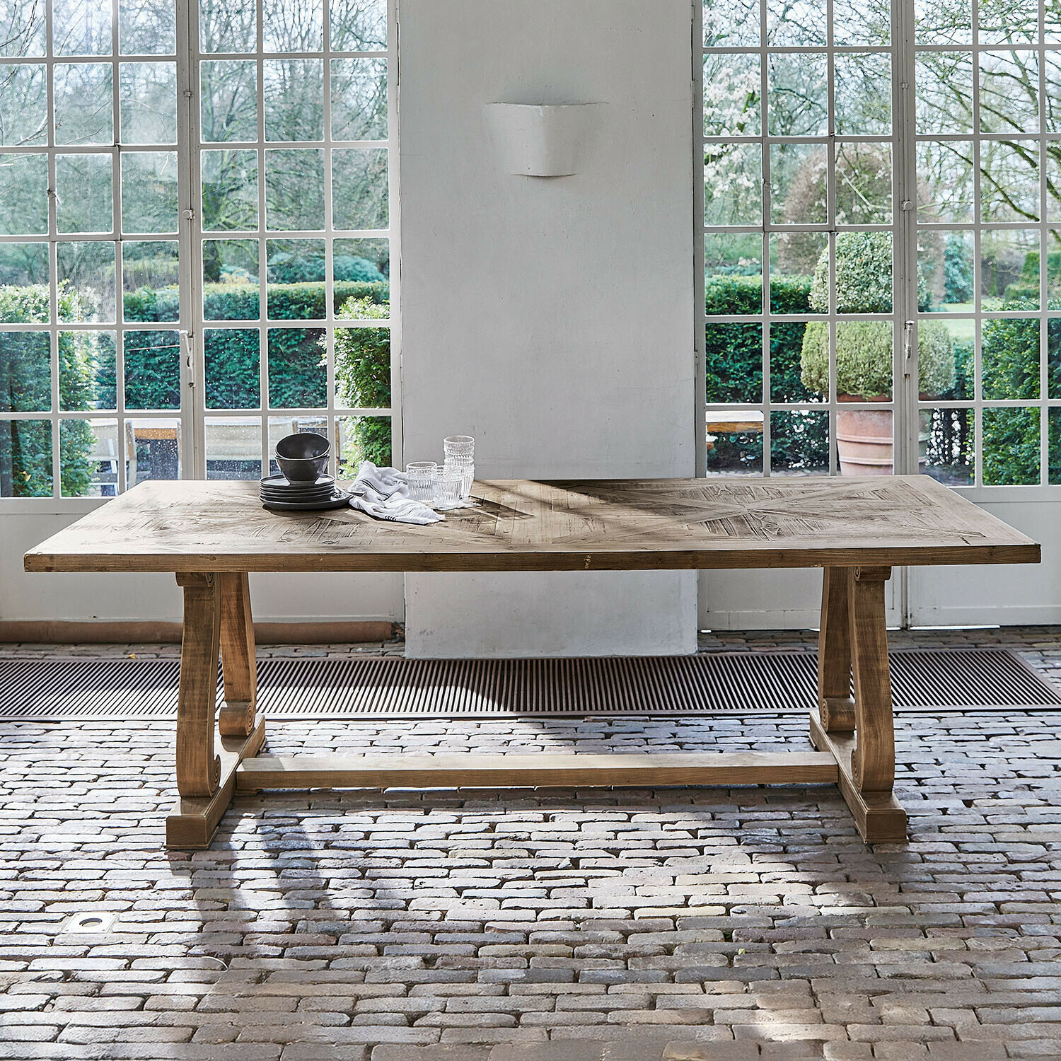 Gran mesa de comedor de madera natural con tablero decorativo frente a grandes ventanas y suelo de ladrillo