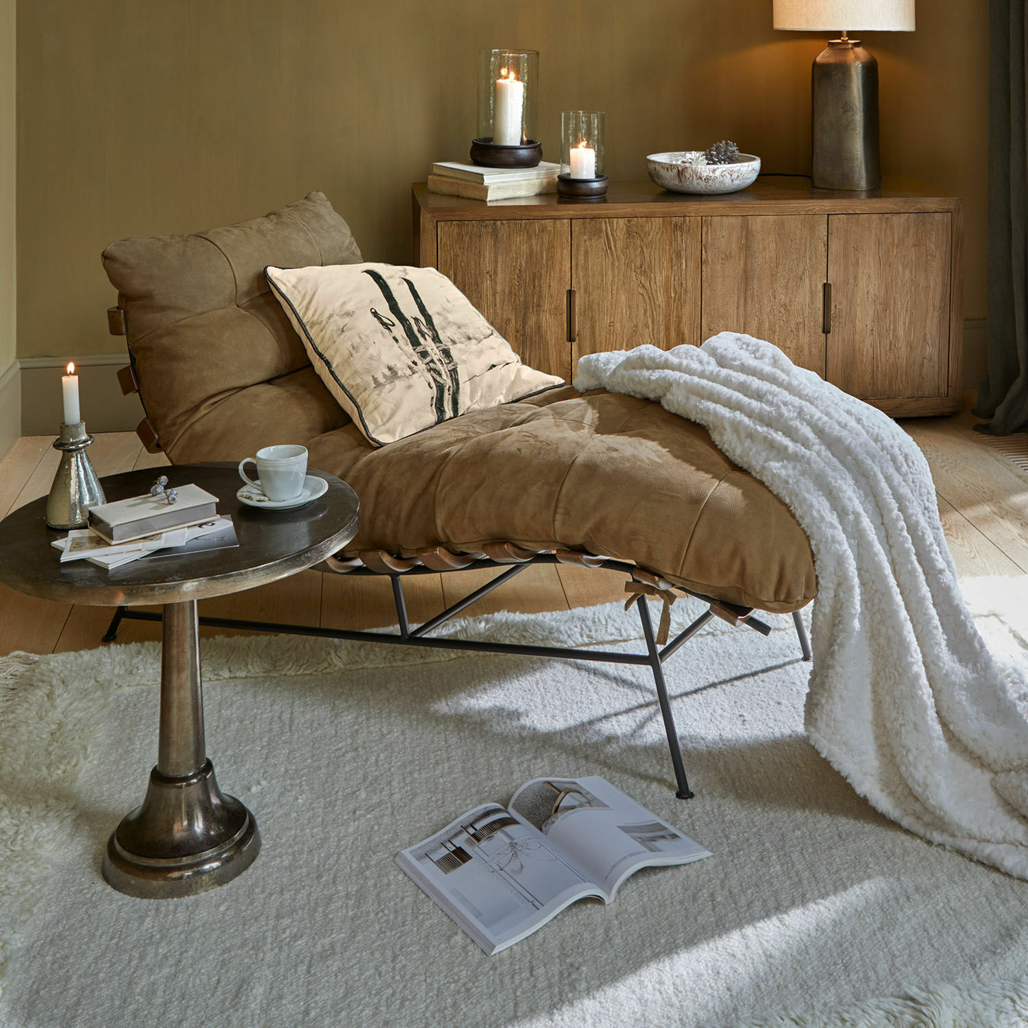 Chaise longue Horraine Sillón de cuero marrón con manta blanca y cojín junto a aparador de madera y mesa auxiliar