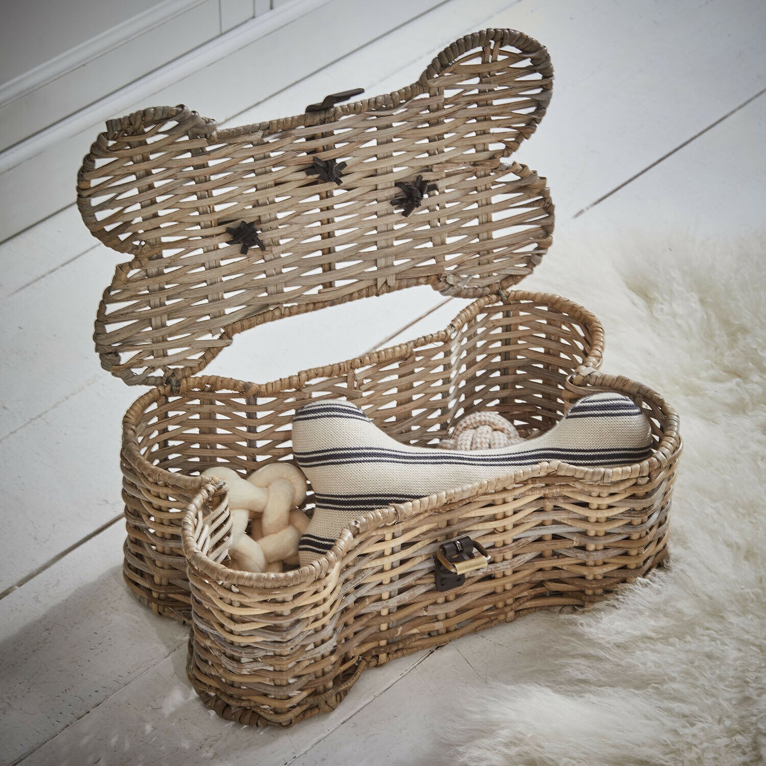 Baúl para mascotas Nuzzlea Cesta de mimbre en forma de hueso con juguetes para perro en color natural sobre suelo blanco