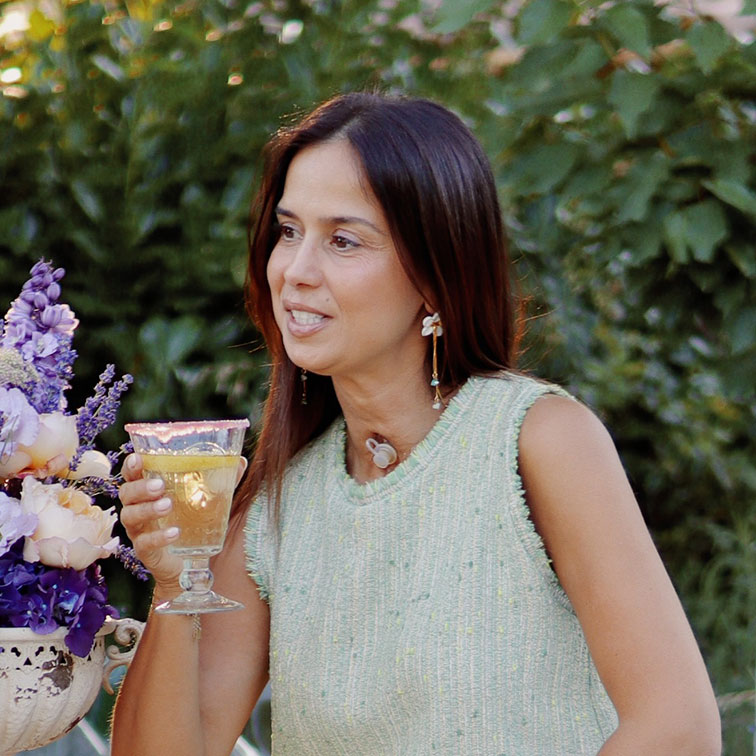 Mujer con suéter verde sin mangas sosteniendo una copa con bebida en un jardín con flores