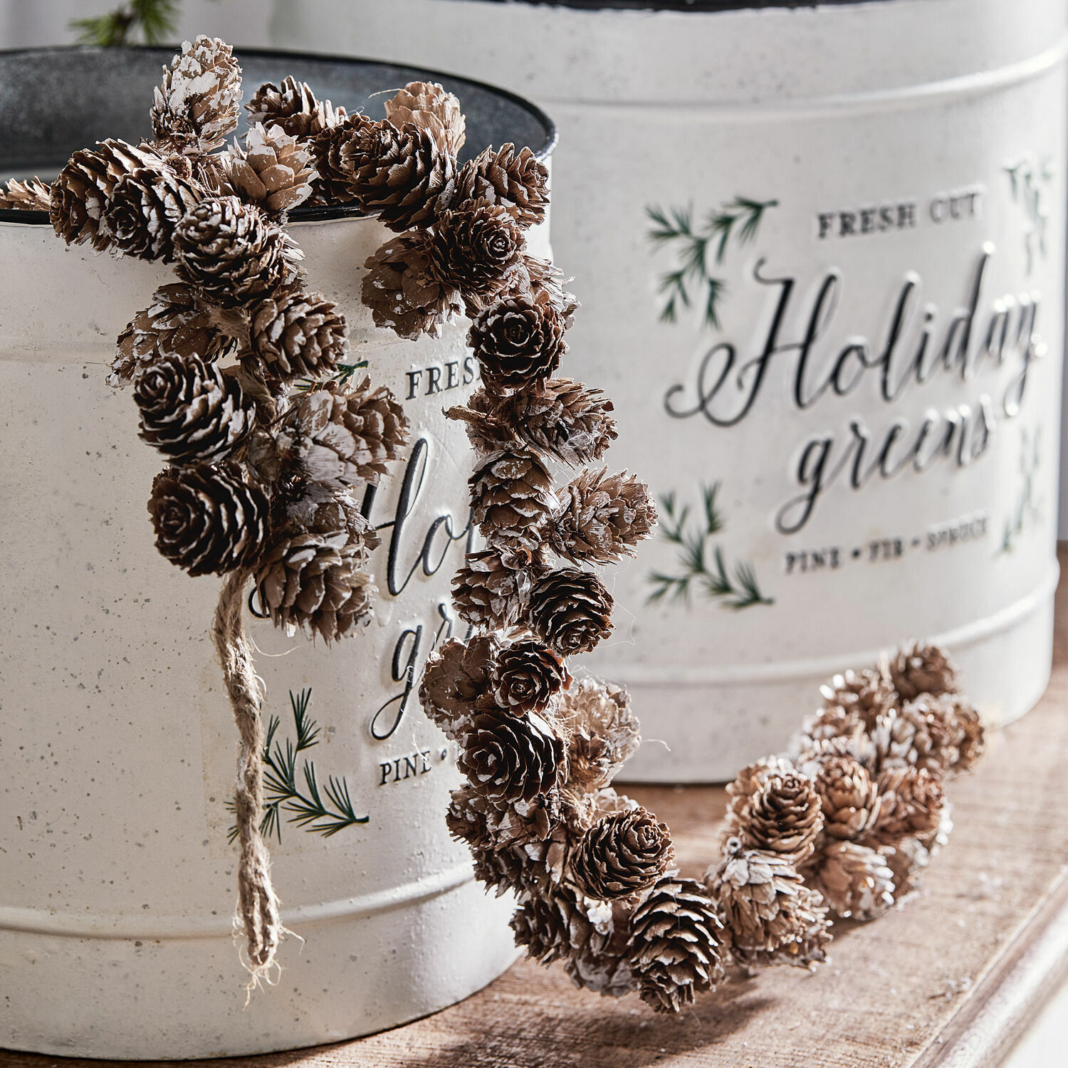 Guirnalda de piñas de abeto Jacci Corona decorativa de piñas en marrón natural con detalles blancos frente a recipientes metálicos blancos