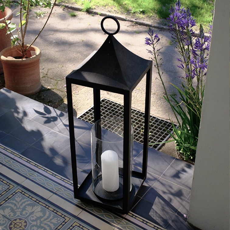 Farol negro de metal con vela blanca y protección de cristal en terraza con baldosas y plantas