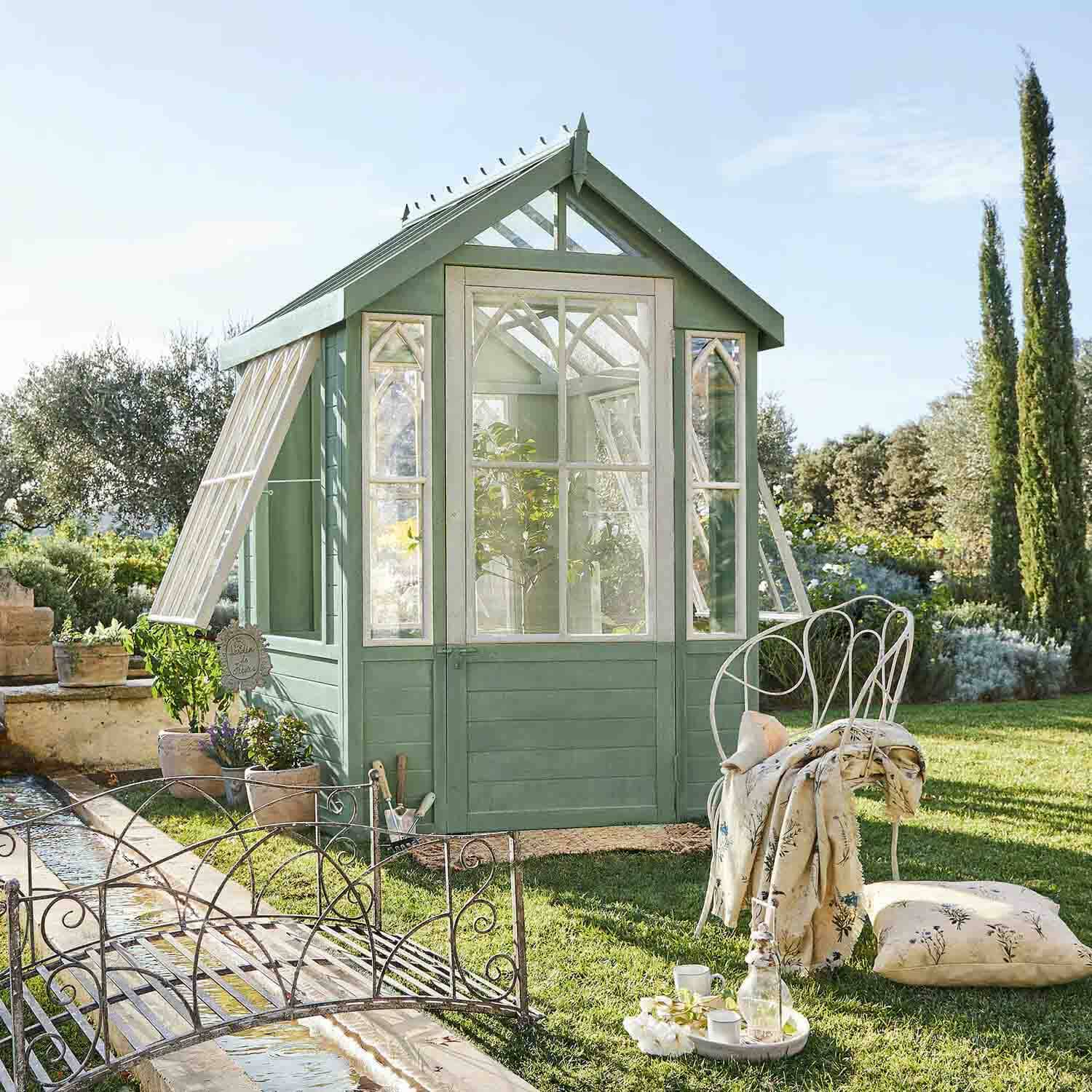 Invernadero de madera verde con marcos de ventana blancos en jardín, silla de metal con cojín al lado
