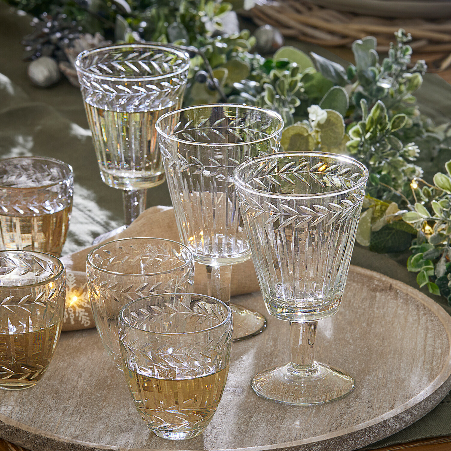 Set de 12 vasos Padiès Conjunto de vasos y copas de cristal transparente con diseño grabado de hojas sobre bandeja de madera