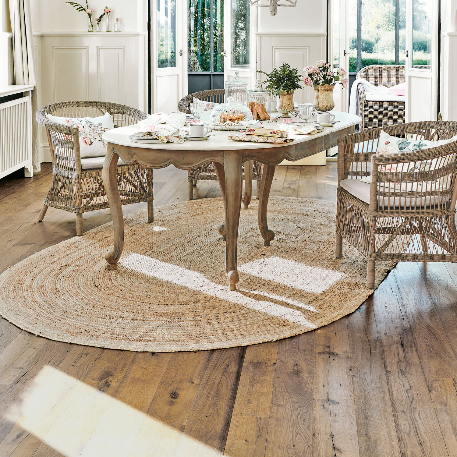 Mesa de madera clara con superficie blanca sobre alfombra redonda de yute en sala luminosa con sillas de mimbre