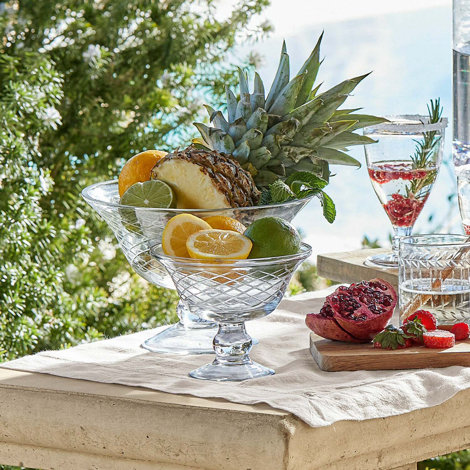 Set de 2 cuencos Kadence Cuencos de vidrio con frutas cítricas y piña sobre mesa clara al aire libre