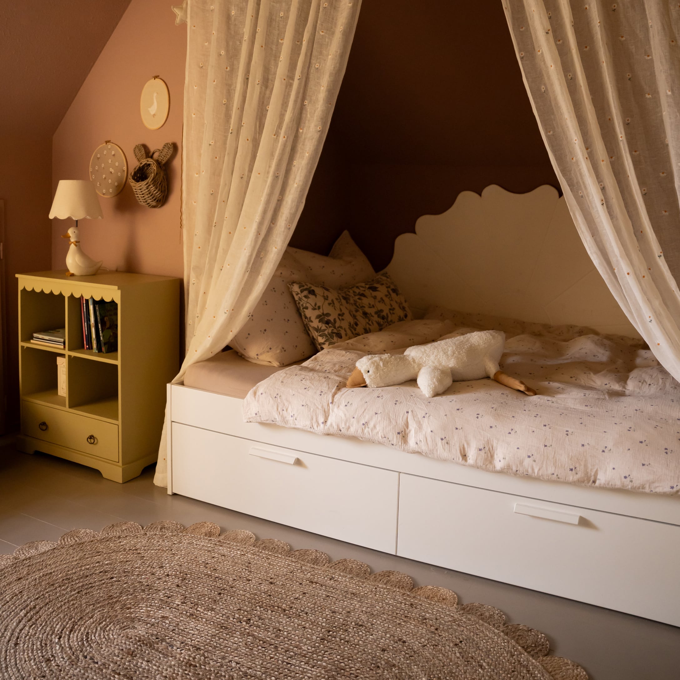 Cama infantil blanca con cortinas y peluche de ganso sobre ropa de cama con estampado floral
