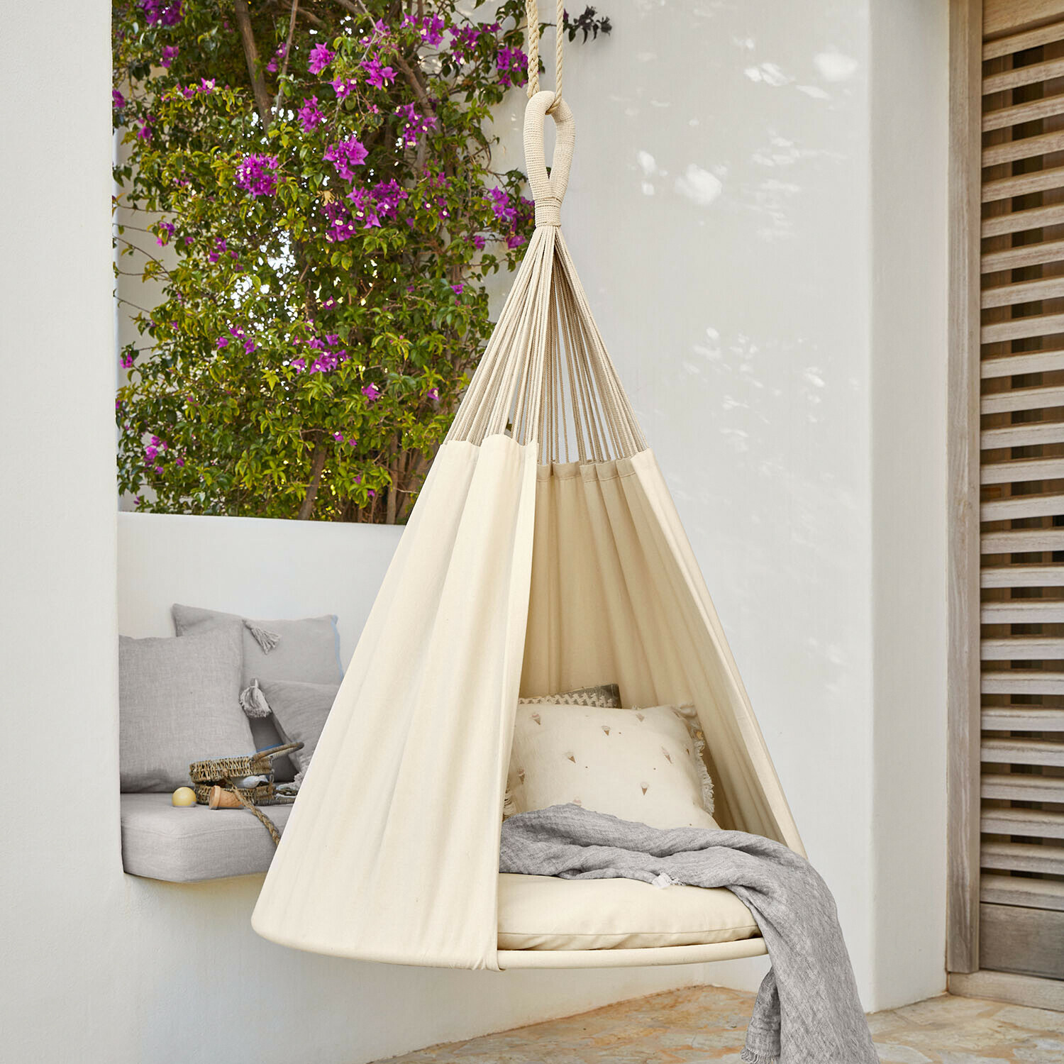 Hamaca colgante beige de algodón con cojines y manta gris en terraza con flores