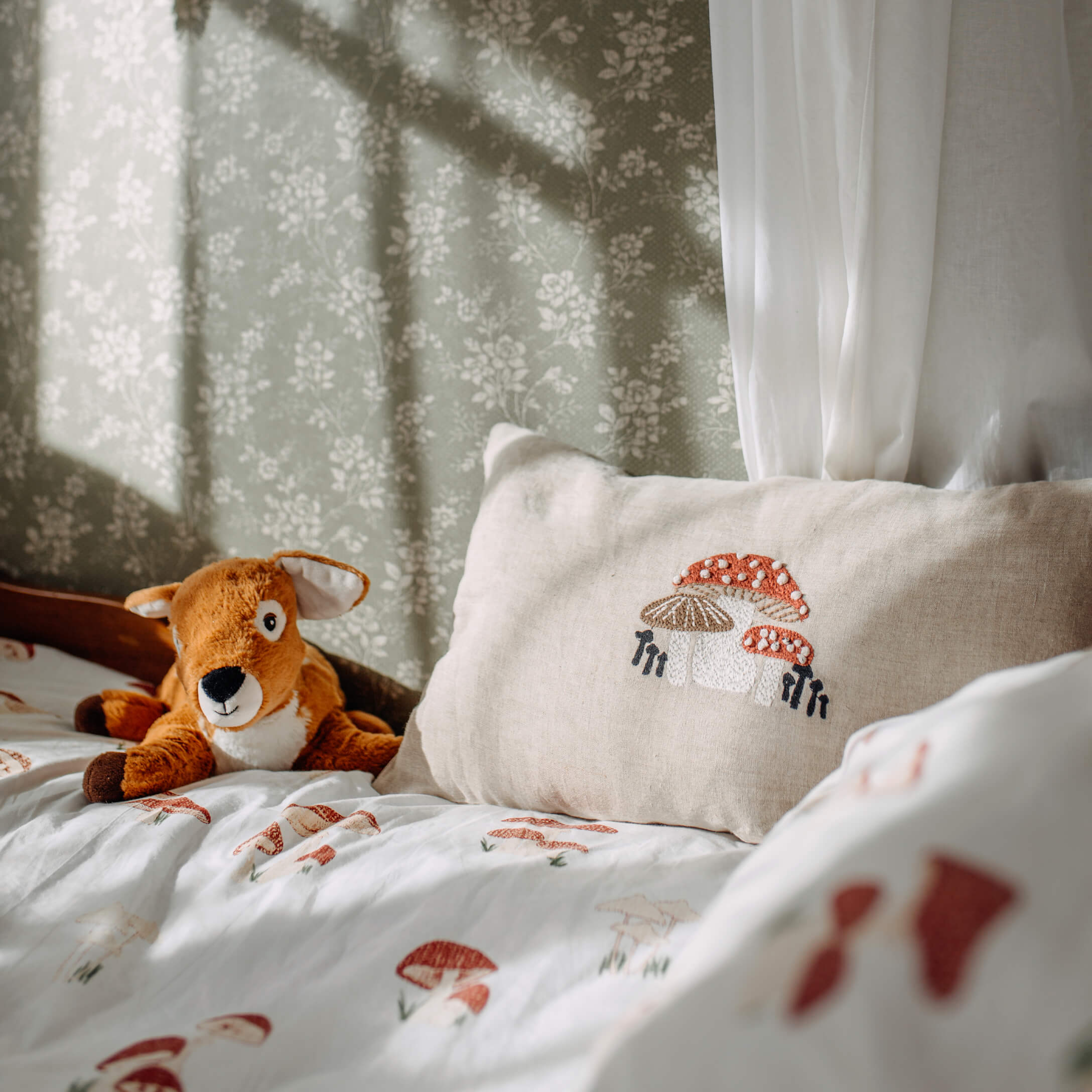 Cojín de lino beige con bordado de setas sobre cama con ropa de cama de setas y peluche de ciervo