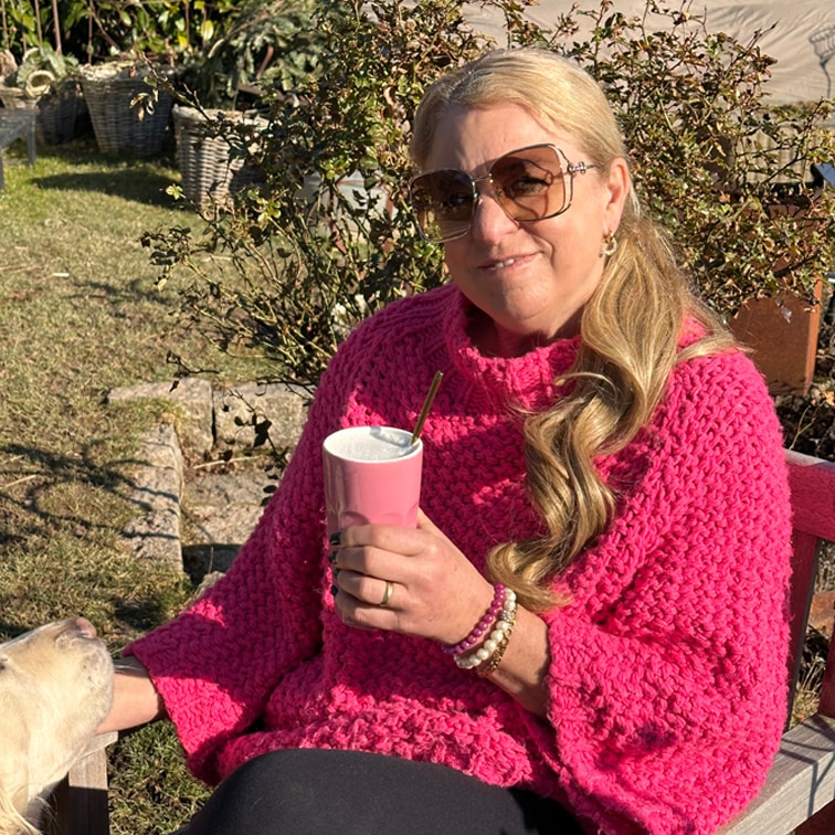 Mujer con suéter grueso rosa sosteniendo taza rosa al aire libre en día soleado con perro al lado