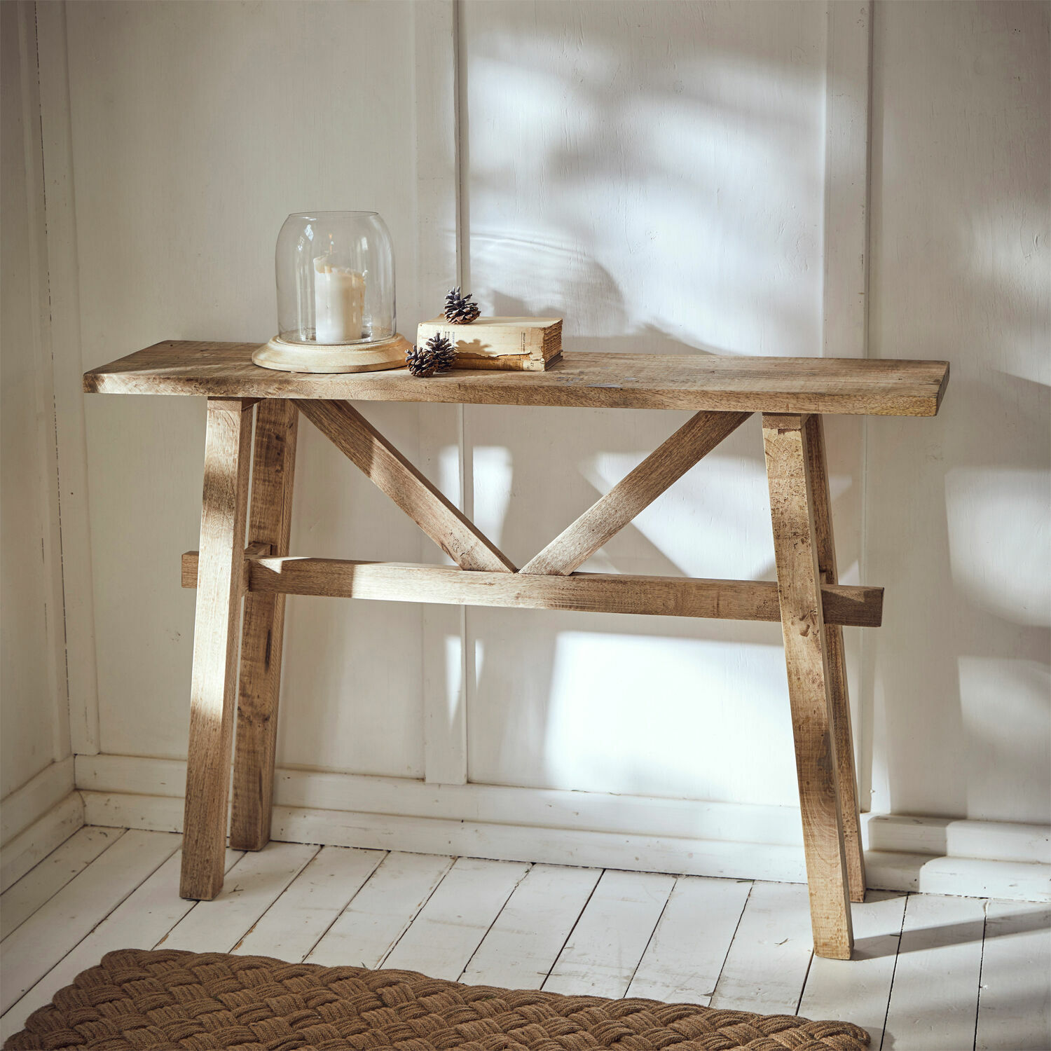 Consola Nael Mesa consola de madera natural con vela bajo campana de cristal y libro sobre suelo blanco de madera