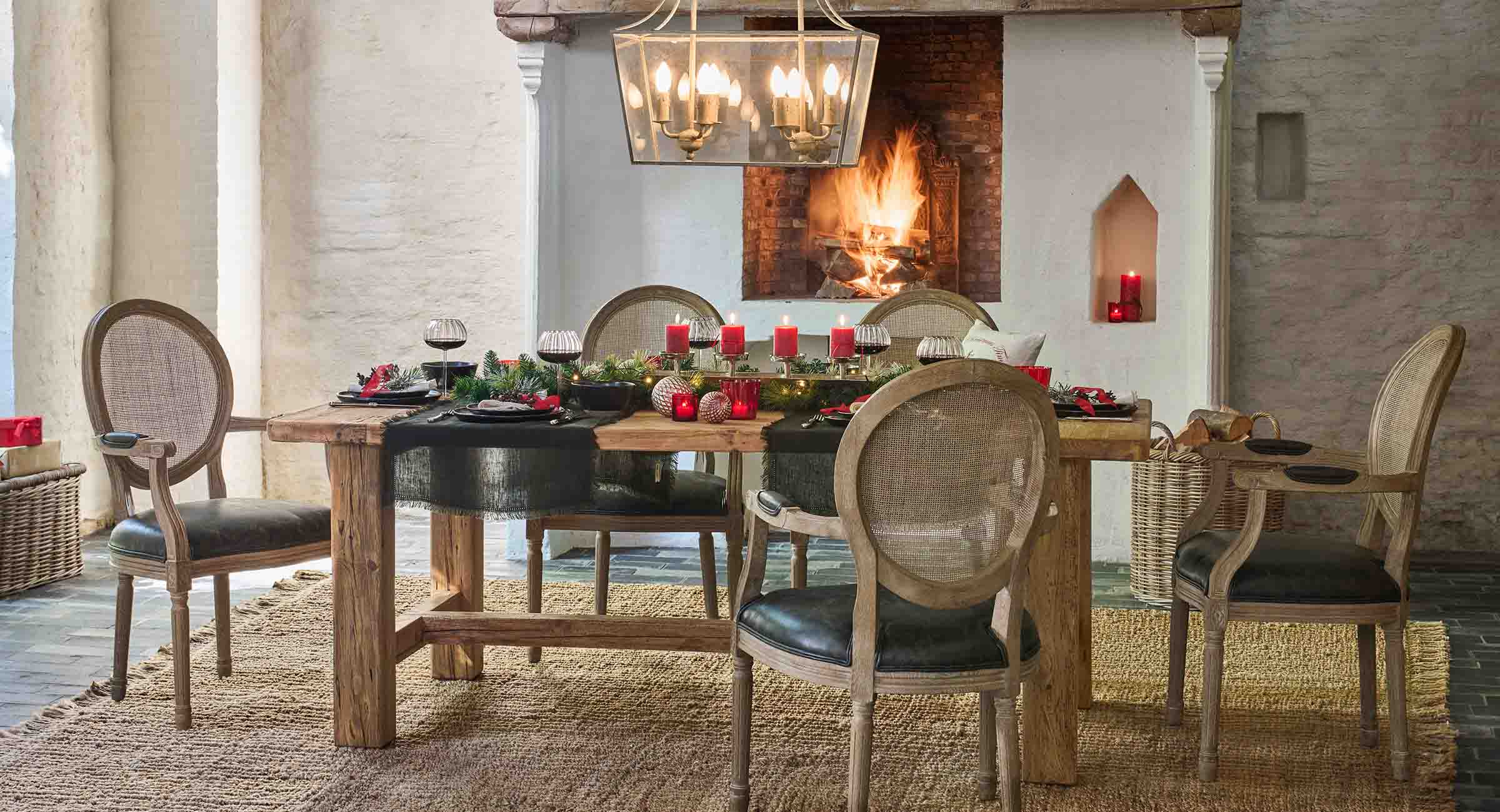 Mesa rústica de madera con decoraciones naturales y velas rojas junto a una chimenea encendida