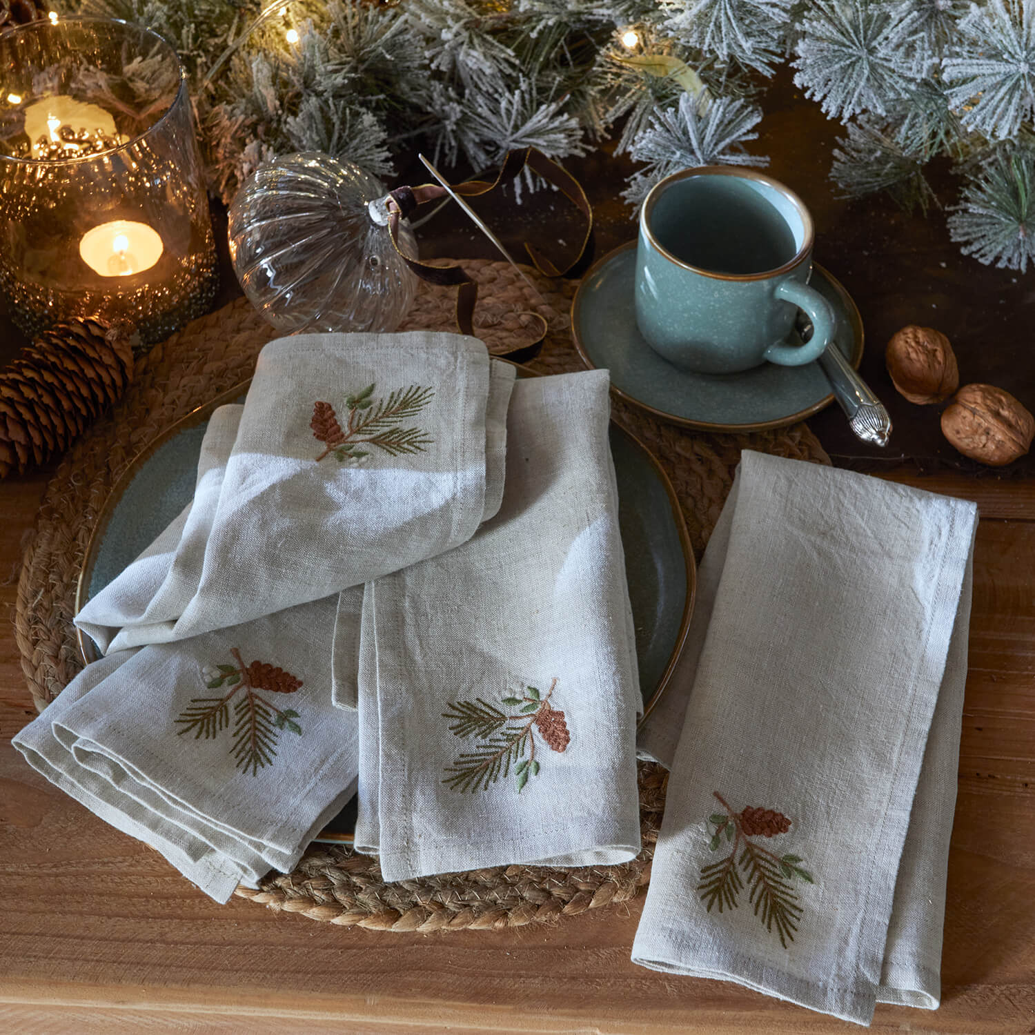 Servilletas de lino bordadas con piñas sobre mesa rústica con taza verde y decoración navideña