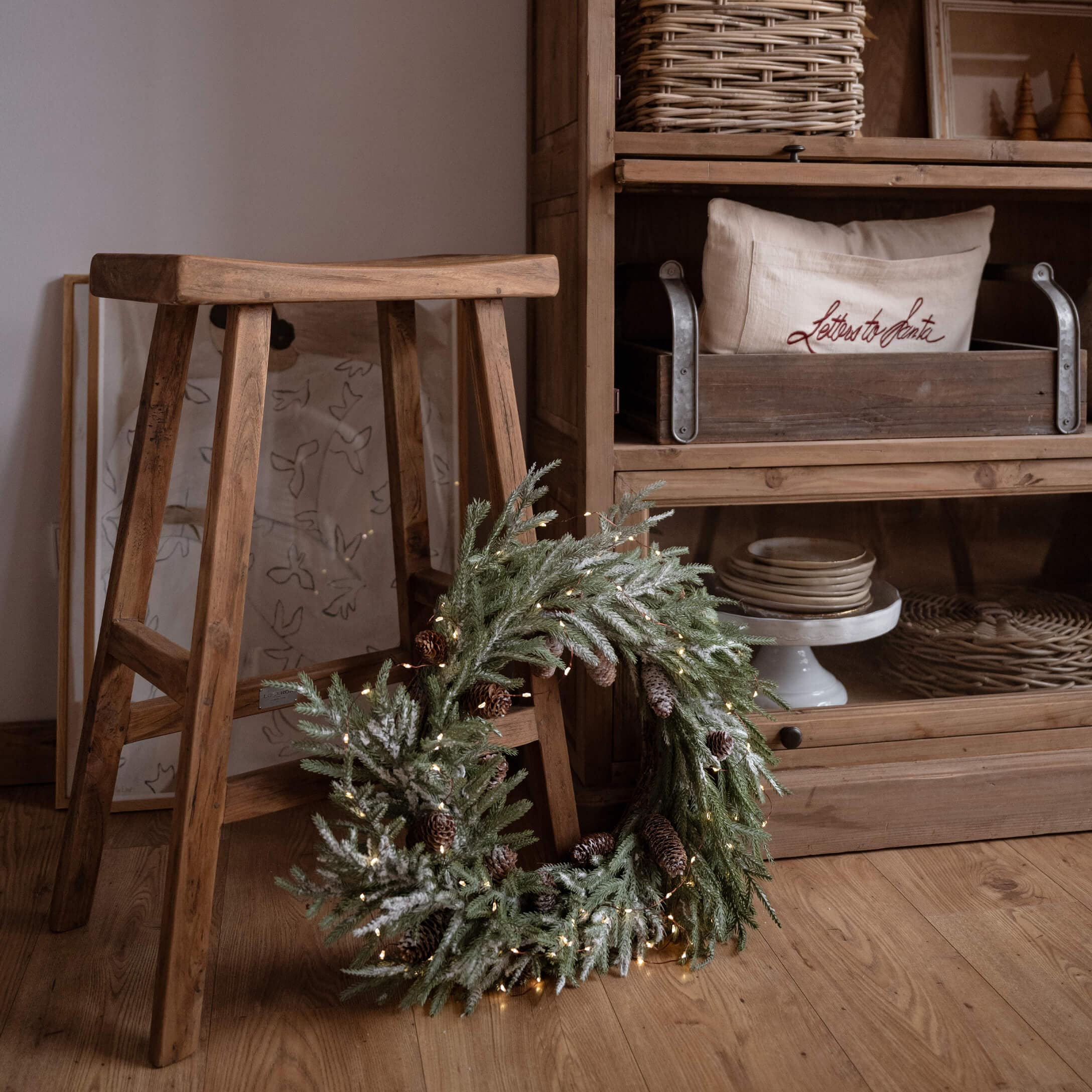 Corona navideña decorativa con ramas verdes y piñas sobre suelo de madera junto a un taburete