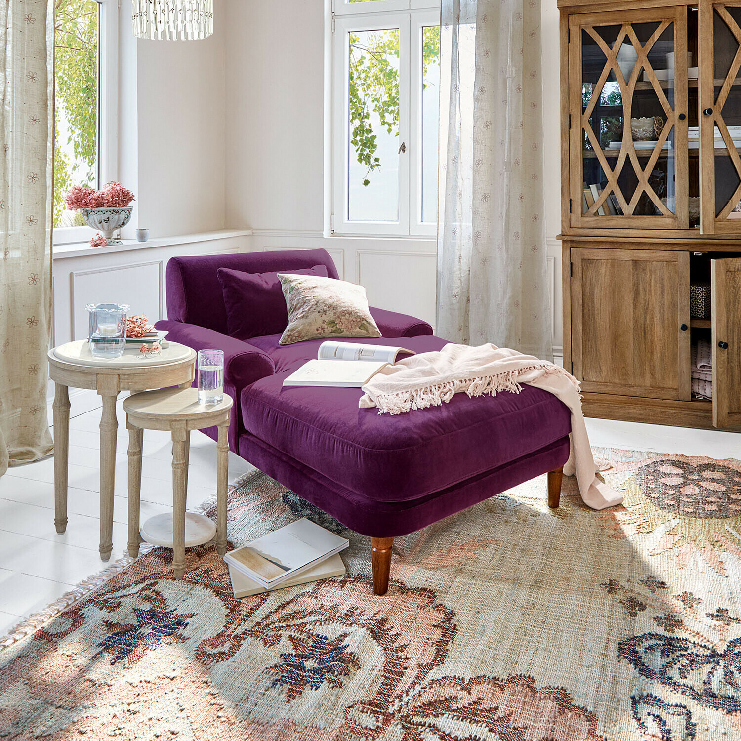 Chaise longue Jouvrelle Chaise longue de terciopelo morado en salón luminoso con armario de madera y mesas redondas