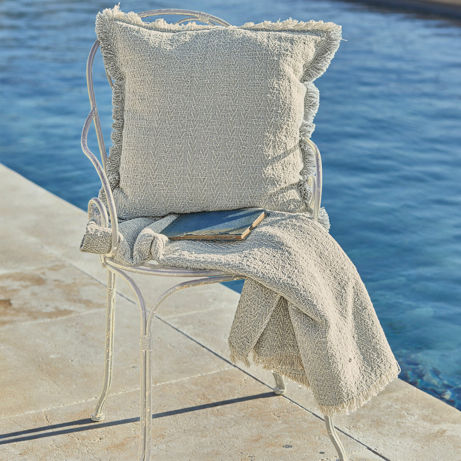 Cojín beige y manta con flecos sobre silla metálica junto al agua, decoración interior y exterior