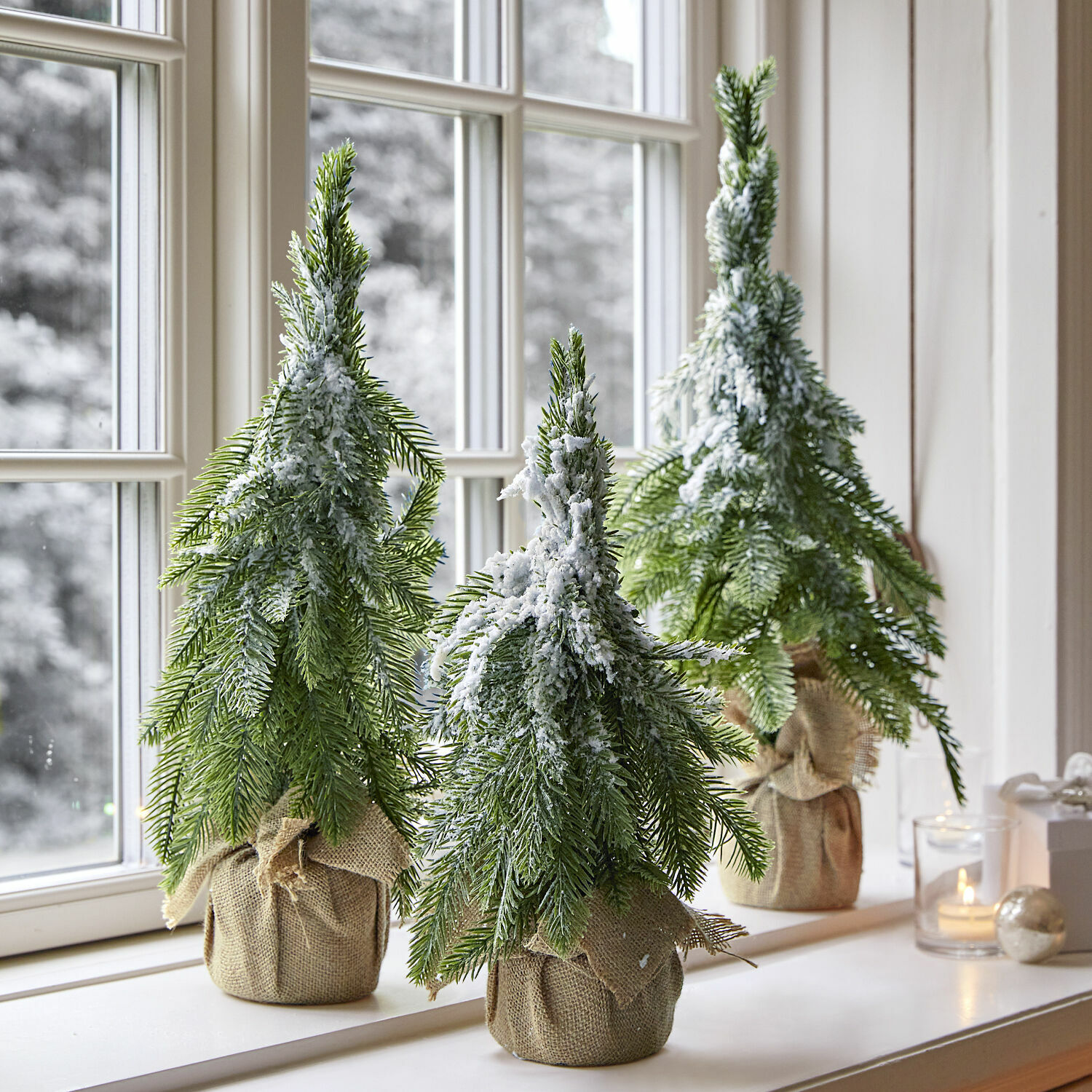 Set de 3 árboles decorativos Selitor Tres árboles de Navidad artificiales con ramas verdes y nieve falsa en base de arpillera sobre el alféizar