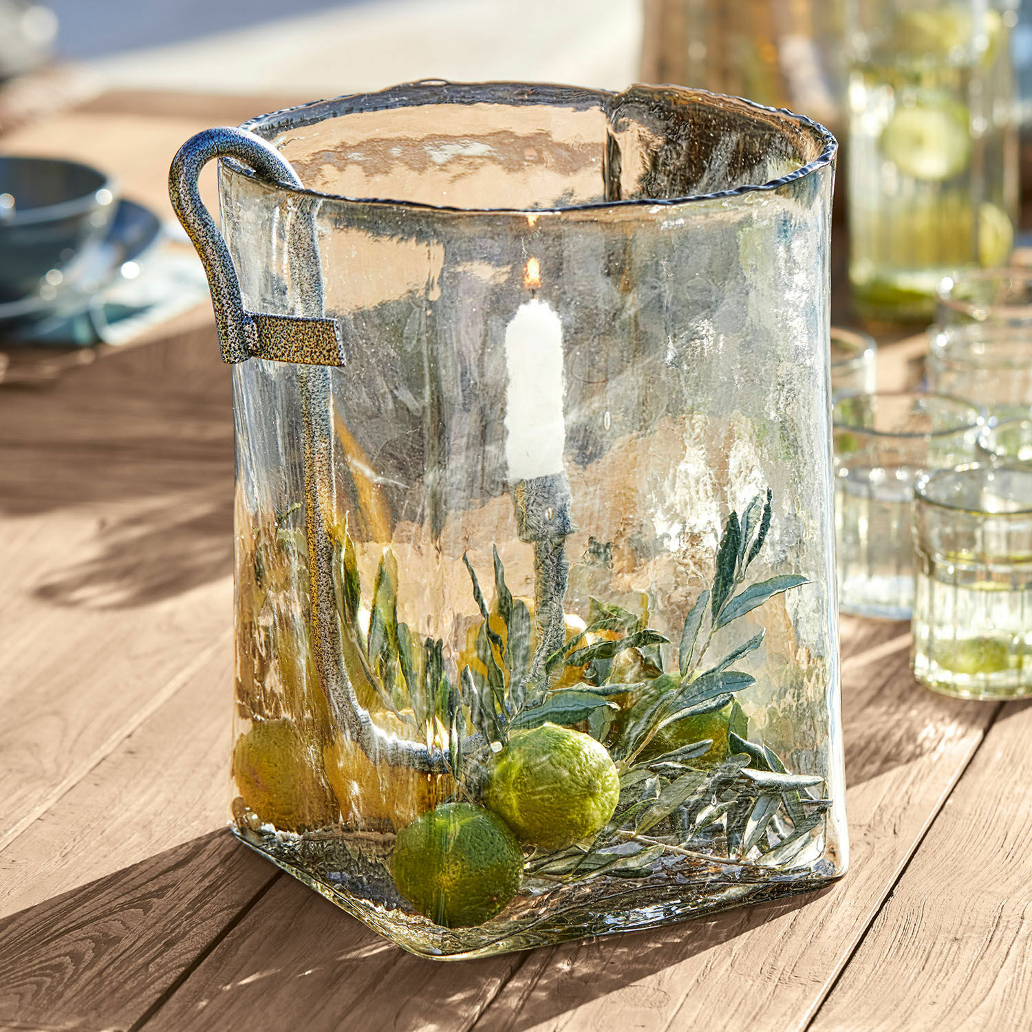 Farol de vidrio con asa metálica y decoración de hojas verdes y limas sobre mesa de madera