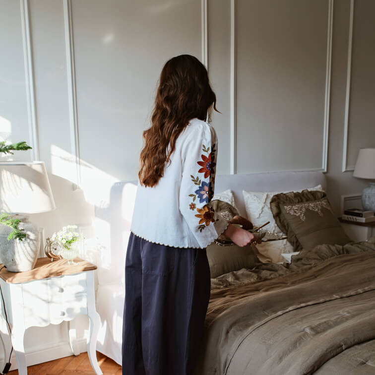 Mujer con blusa blanca bordada de colores en dormitorio con ropa de cama marrón y lámpara sobre mesita