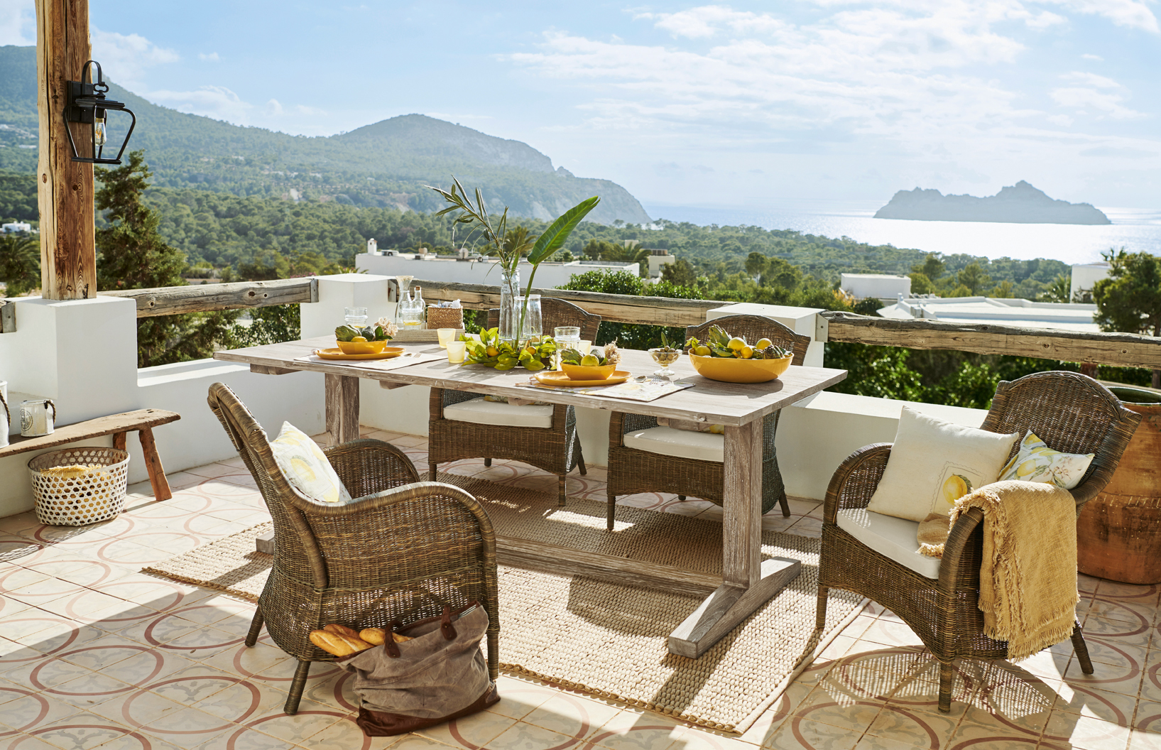 Terraza con muebles de ratán y mesa de madera clara con vistas al mar y montañas, estilo mediterráneo