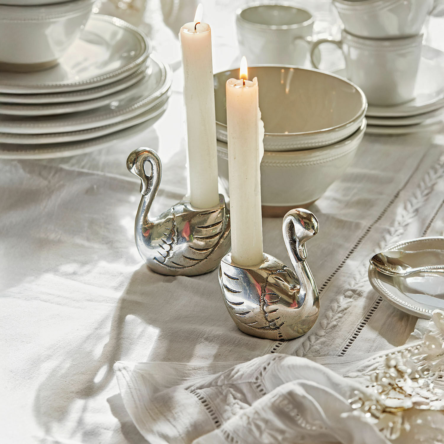 Set de 2 candeleros Zaymara Par de candelabros plateados en forma de cisne sobre mantel blanco con vajilla blanca