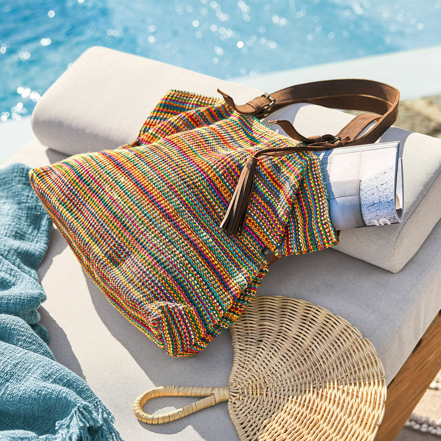 Bolso Gillian Bolso tejido multicolor con correa marrón sobre tumbona clara junto a la piscina, accesorio veraniego