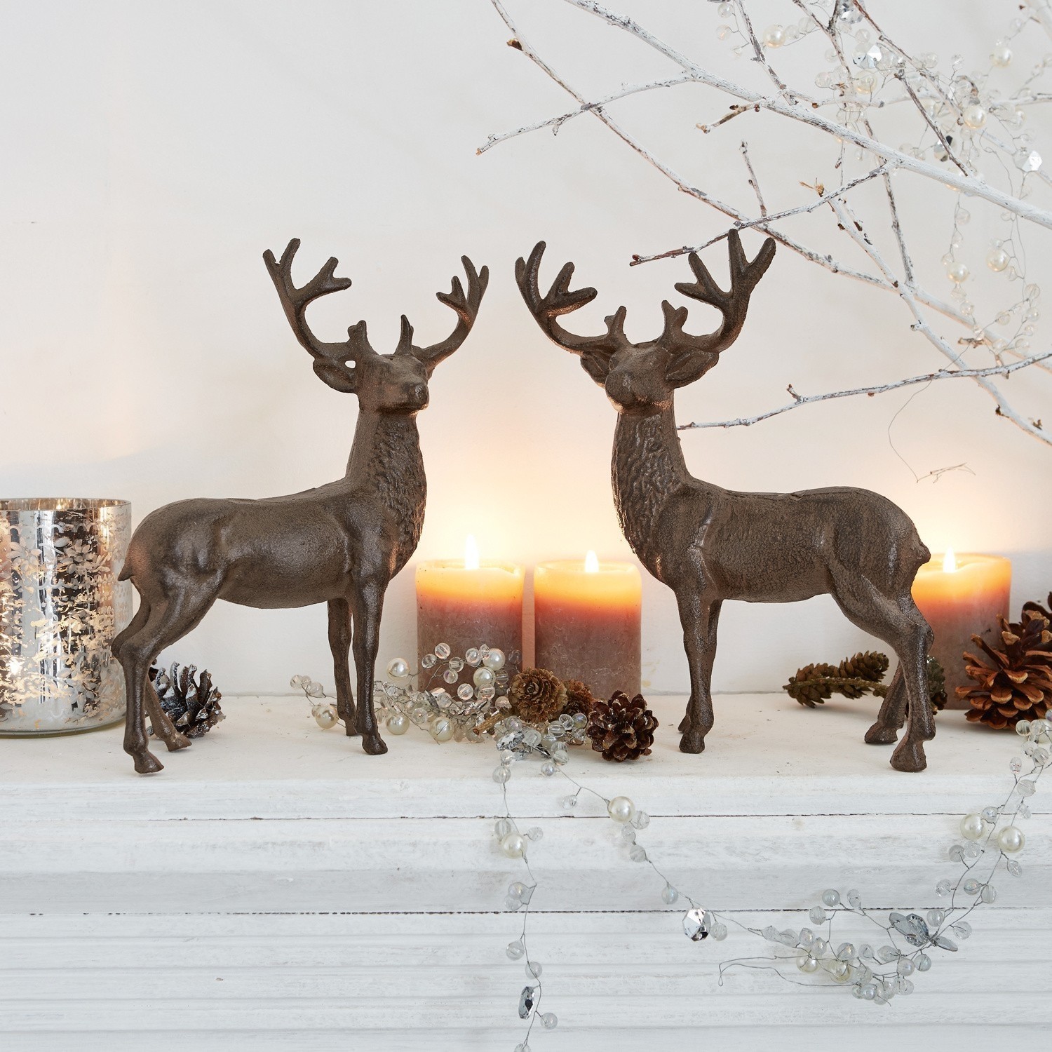 Set de 2 ciervos decorativos Friends Dos figuras de ciervos de metal marrón sobre estante blanco con velas y piñas decorativas