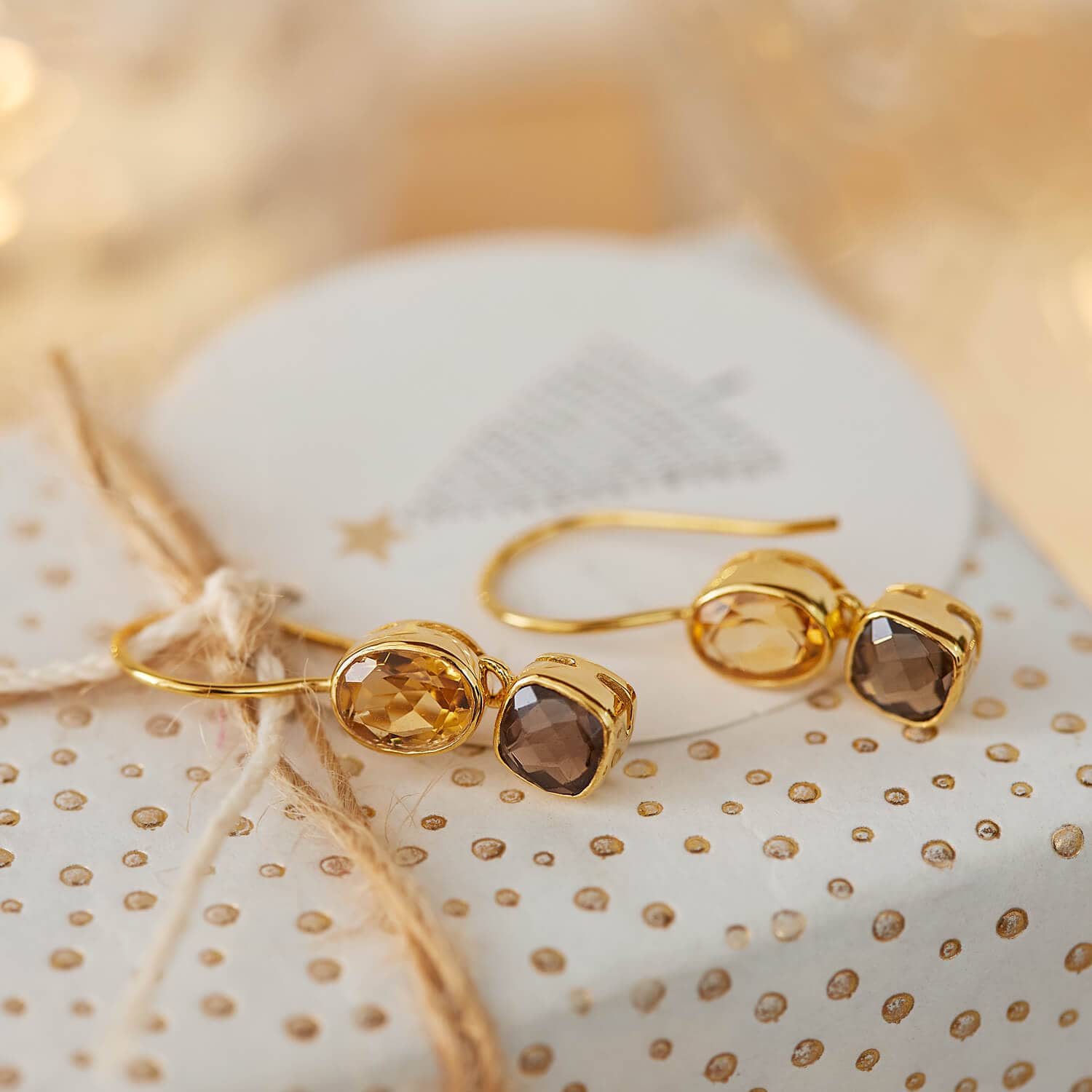 Pendientes dorados con piedras facetadas en tonos marrón y ámbar sobre fondo decorativo festivo
