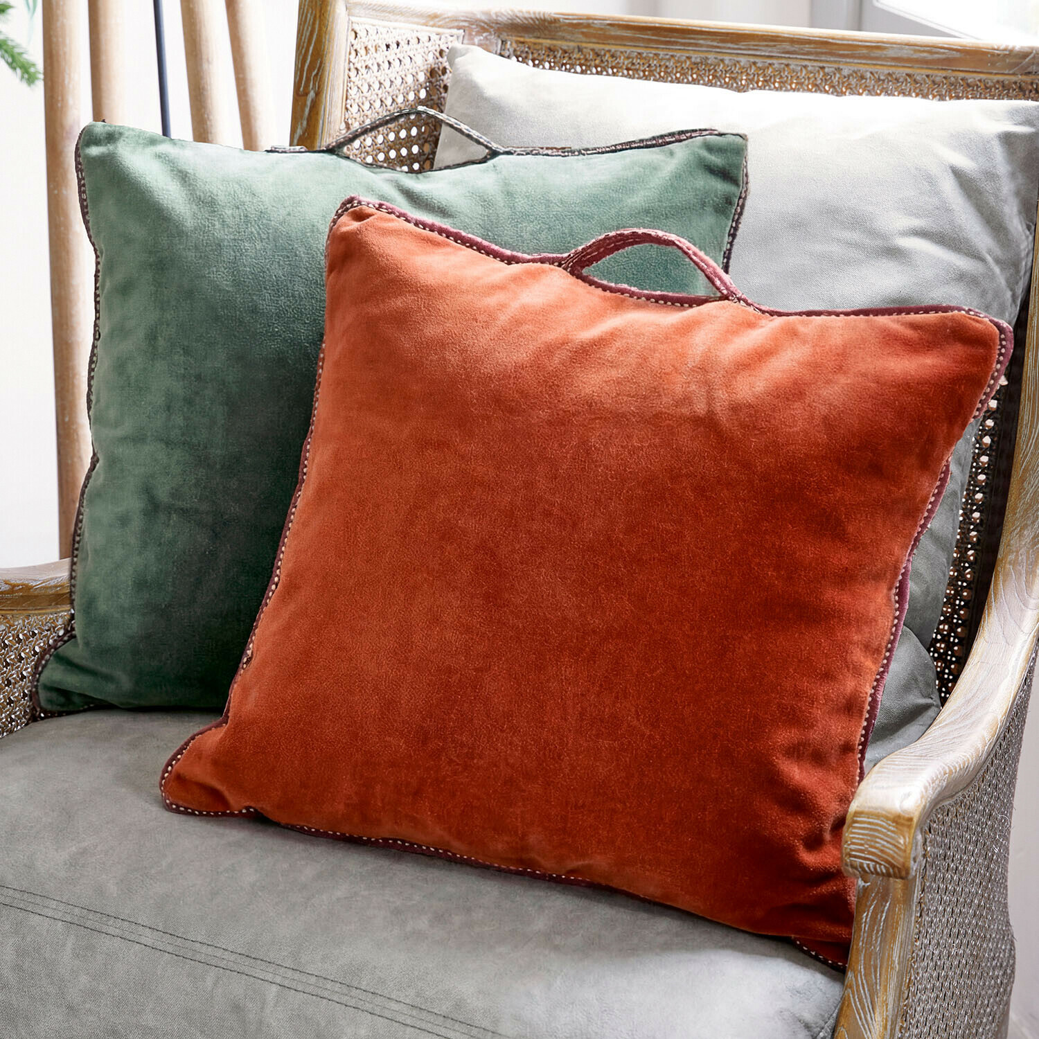 Tres cojines decorativos de terciopelo en verde, naranja y gris sobre un sillón de mimbre para un ambiente acogedor