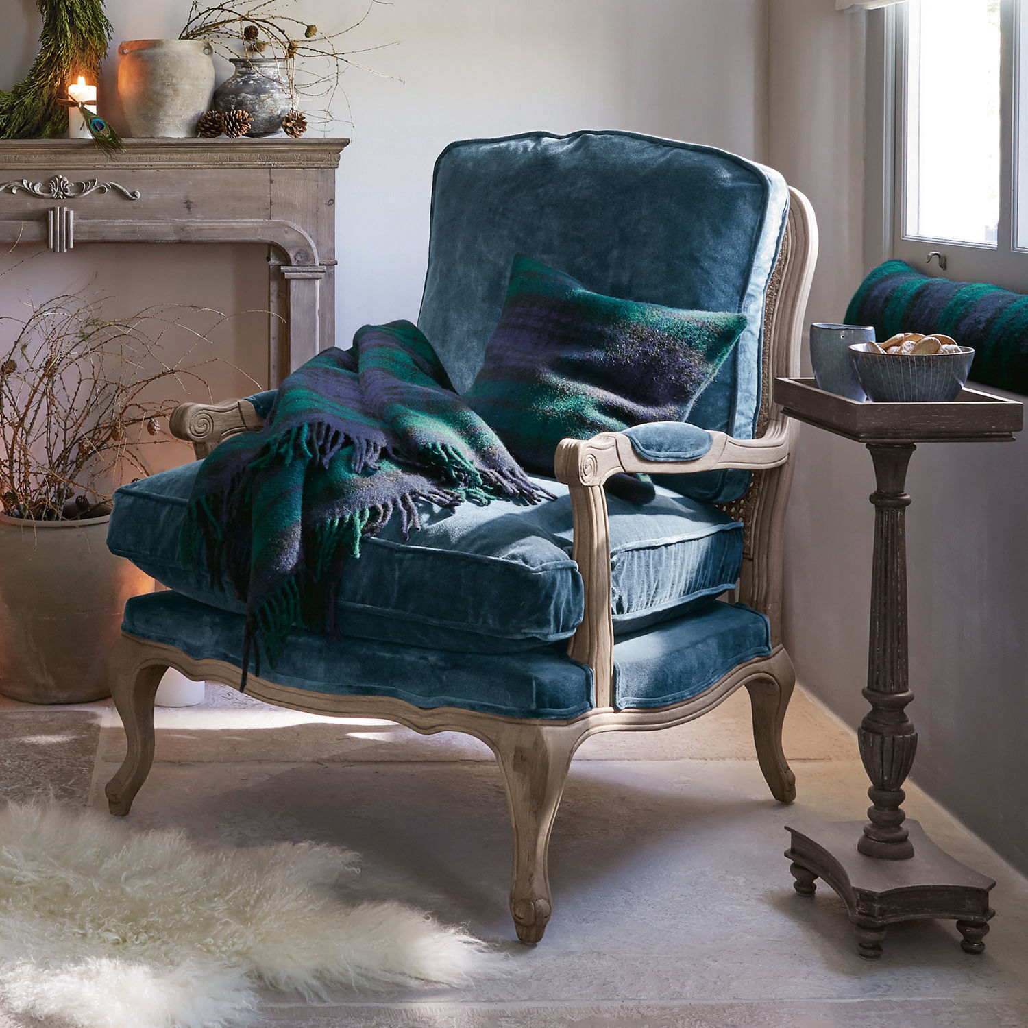 Sillón azul de terciopelo con estructura de madera clara, manta y cojín verde y azul oscuro