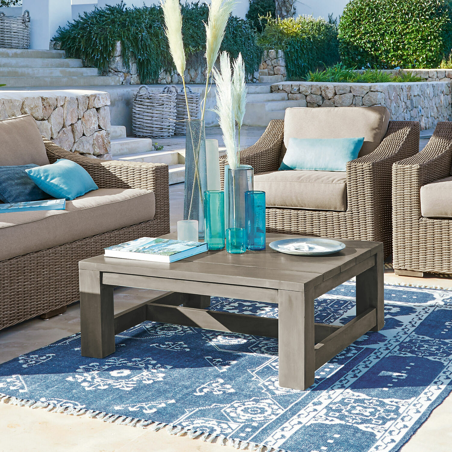Mesa de centro Malden Mesa de centro de madera gris sobre alfombra azul con patrón, rodeada de sillones de mimbre con cojines azules.