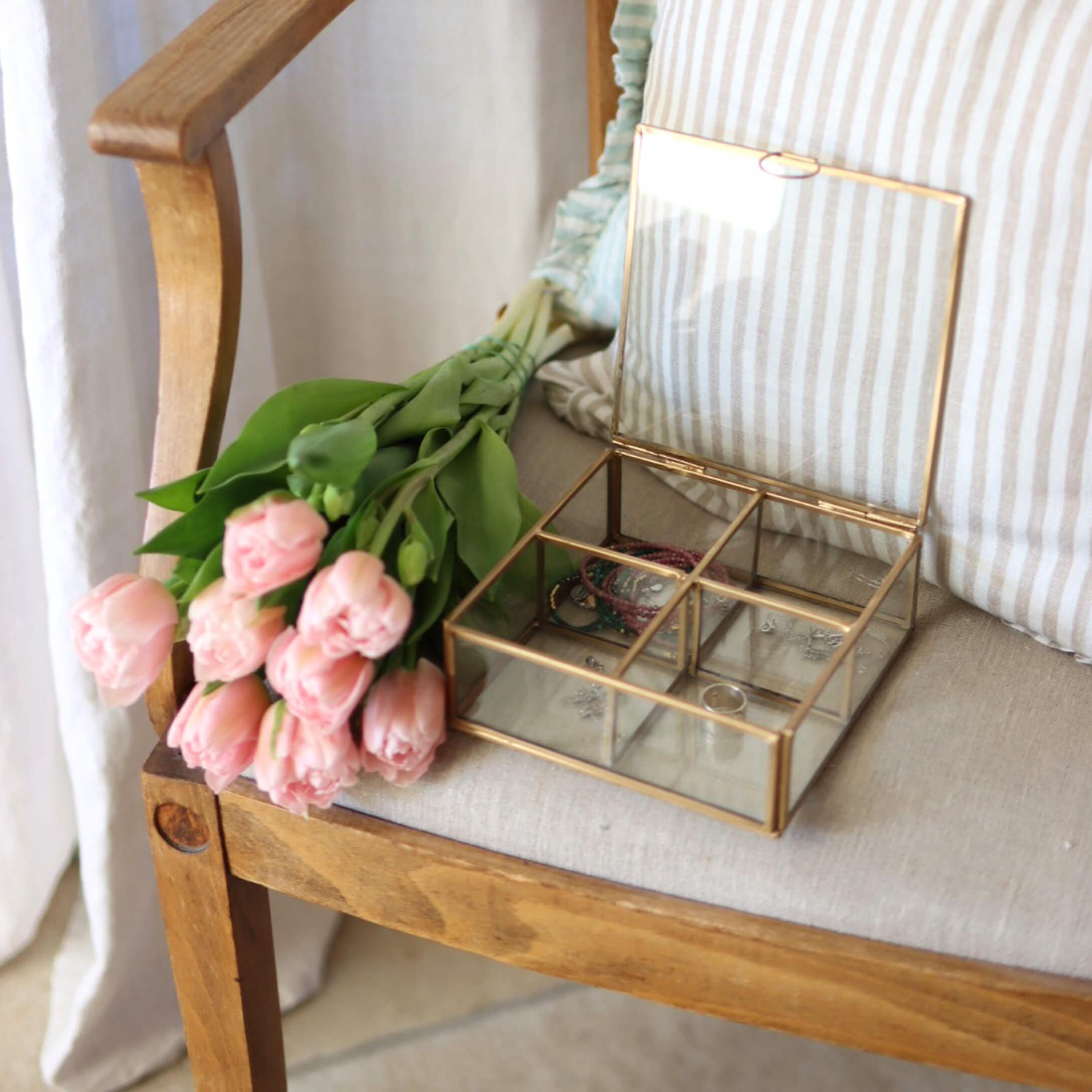 Caja de joyería de vidrio con marco de latón sobre silla de madera junto a tulipanes rosas