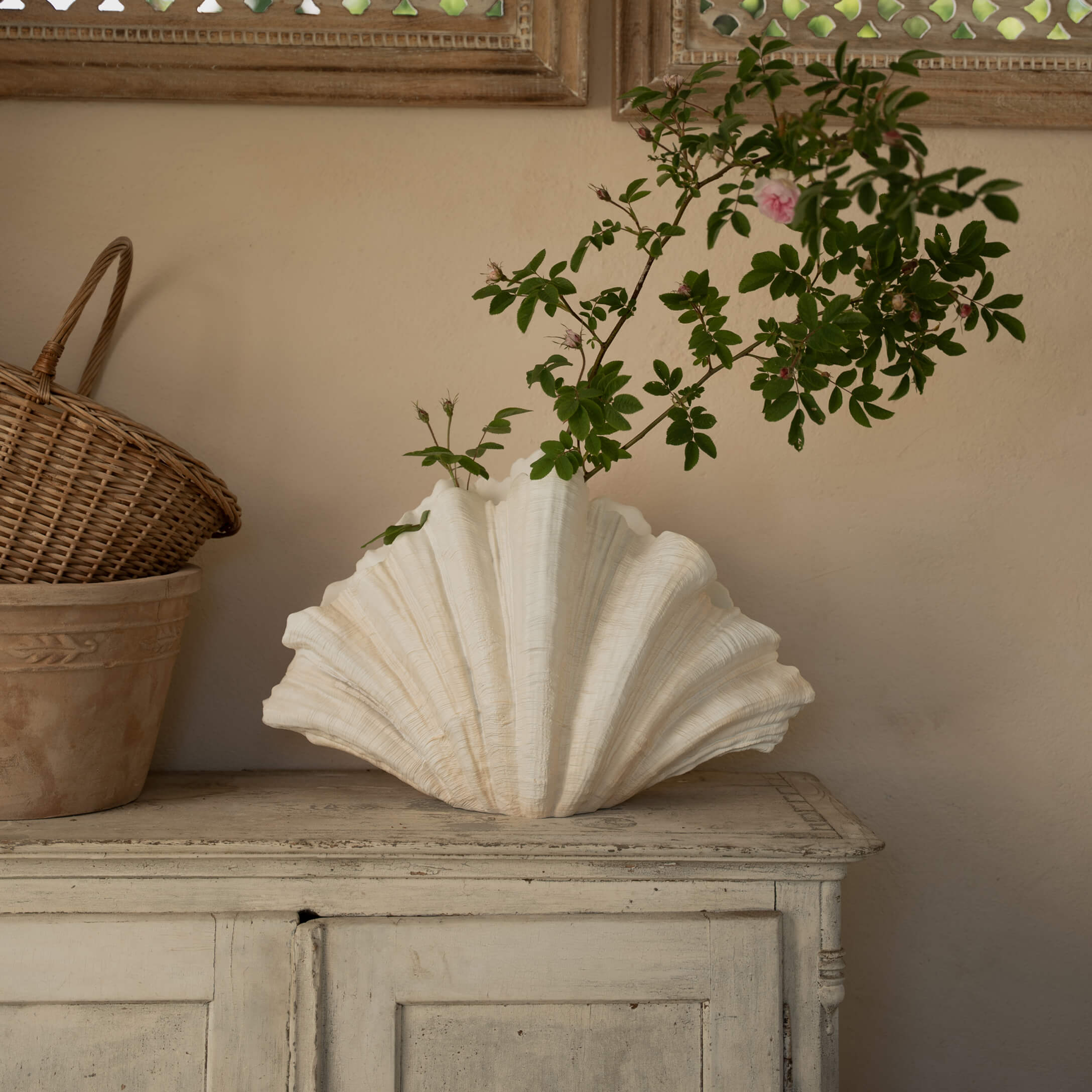 Jarrón de cerámica blanca con forma de concha y ramas verdes sobre cómoda de madera