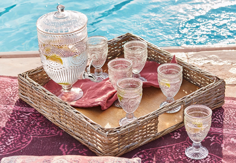 Jarra de vidrio y seis vasos decorados en bandeja de mimbre junto a la piscina, set de bebidas de verano