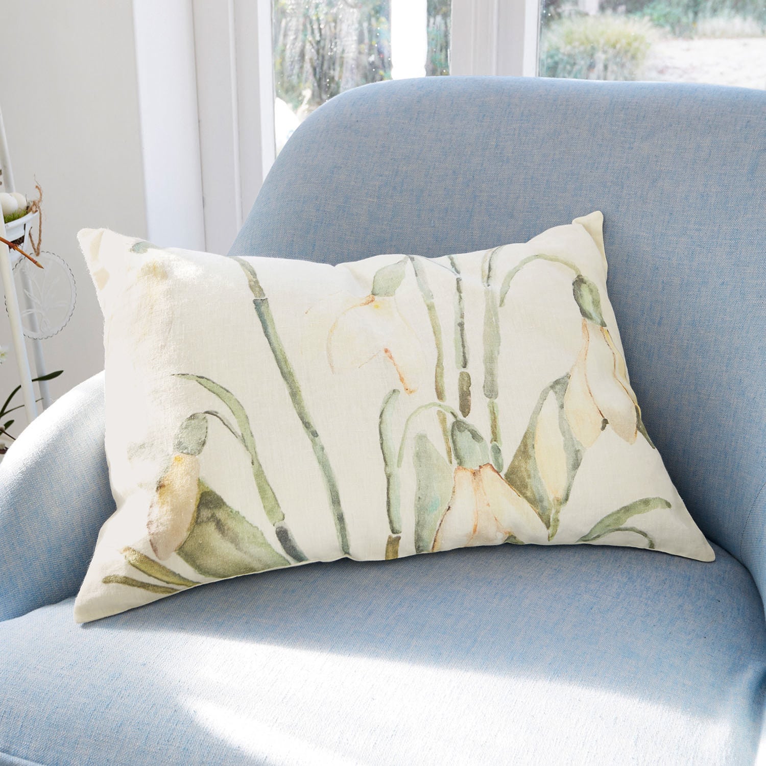 Cojín decorativo rectangular de algodón color crema con estampado floral verde y amarillo sobre sillón azul